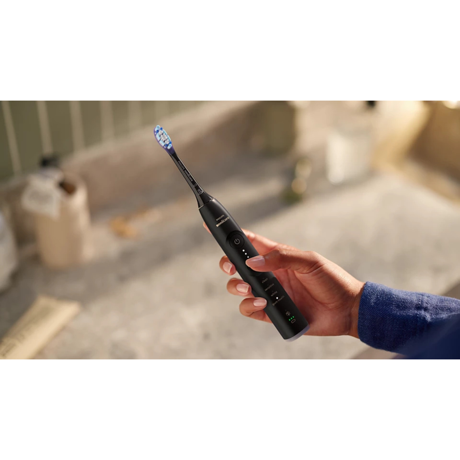 Une brosse à dents électrique noire est tenue par une main au-dessus d'une scène de salle de bain floue.
