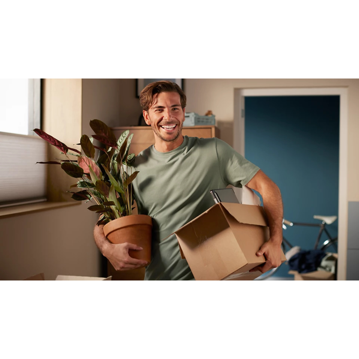 Homme souriant, tenant plante et carton dans une pièce.