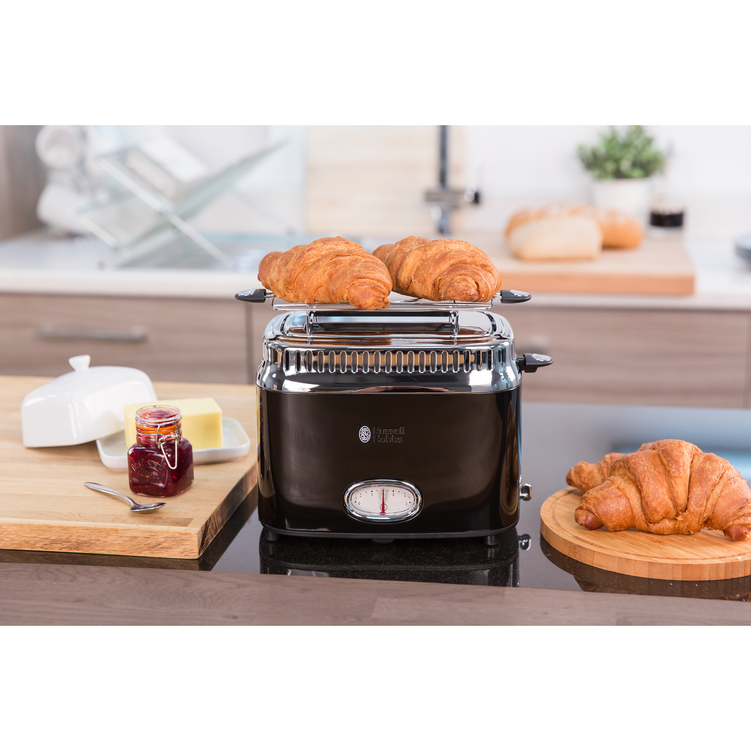 Black toaster with croissants on top, butter and jam, set on a kitchen counter.