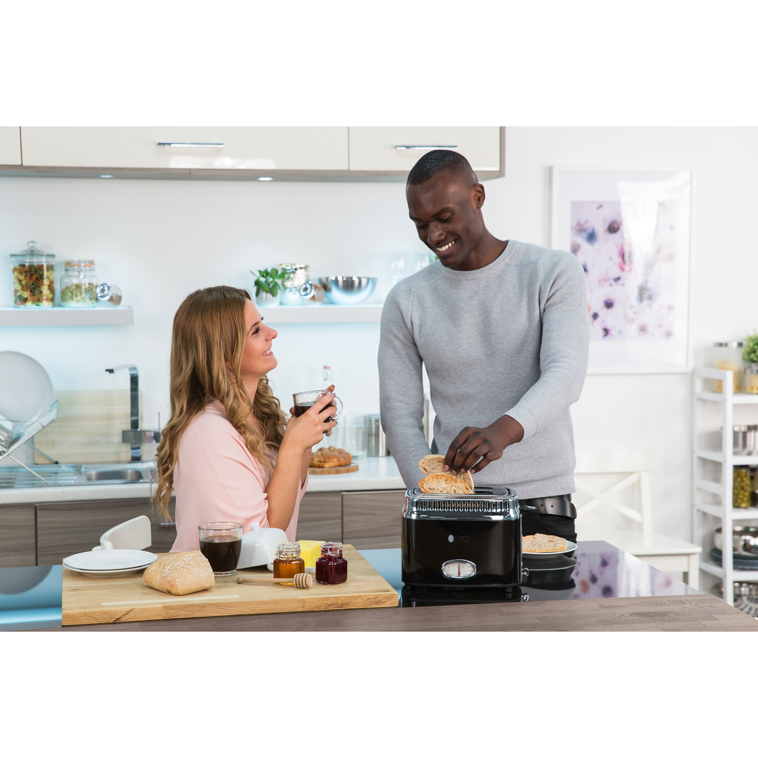 A couple in a kitchen, man toasting bread, woman drinking coffee, with bread and jam on the table.