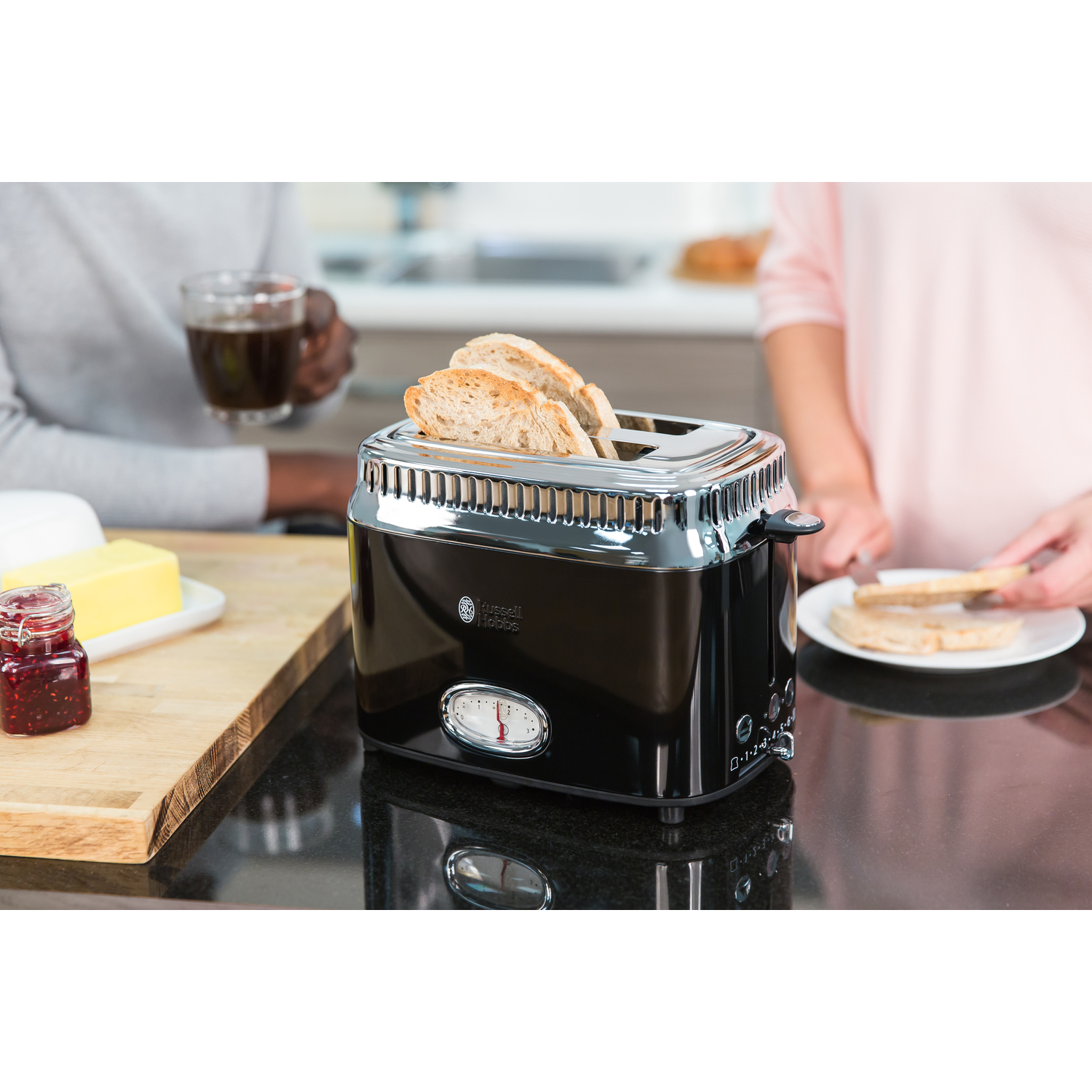 Toaster with bread slices. Two people are present, preparing or eating toast with butter and jam.
