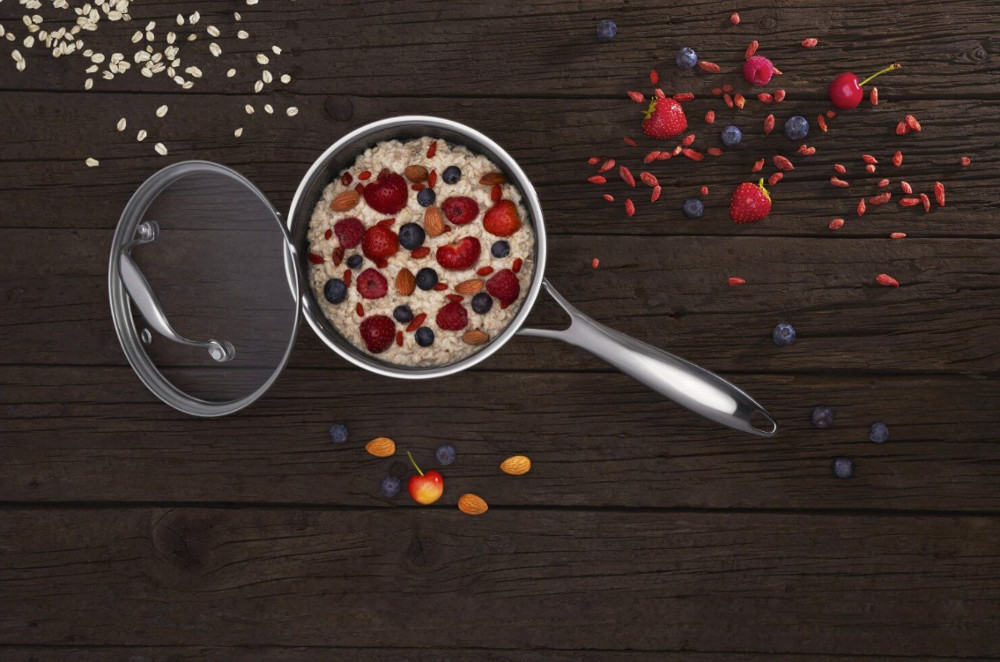 Owsianka w garnku, posypana jagodami i orzechami. Stoi na ciemnej drewnianej powierzchni. Dodatkowe jagody wokoło.