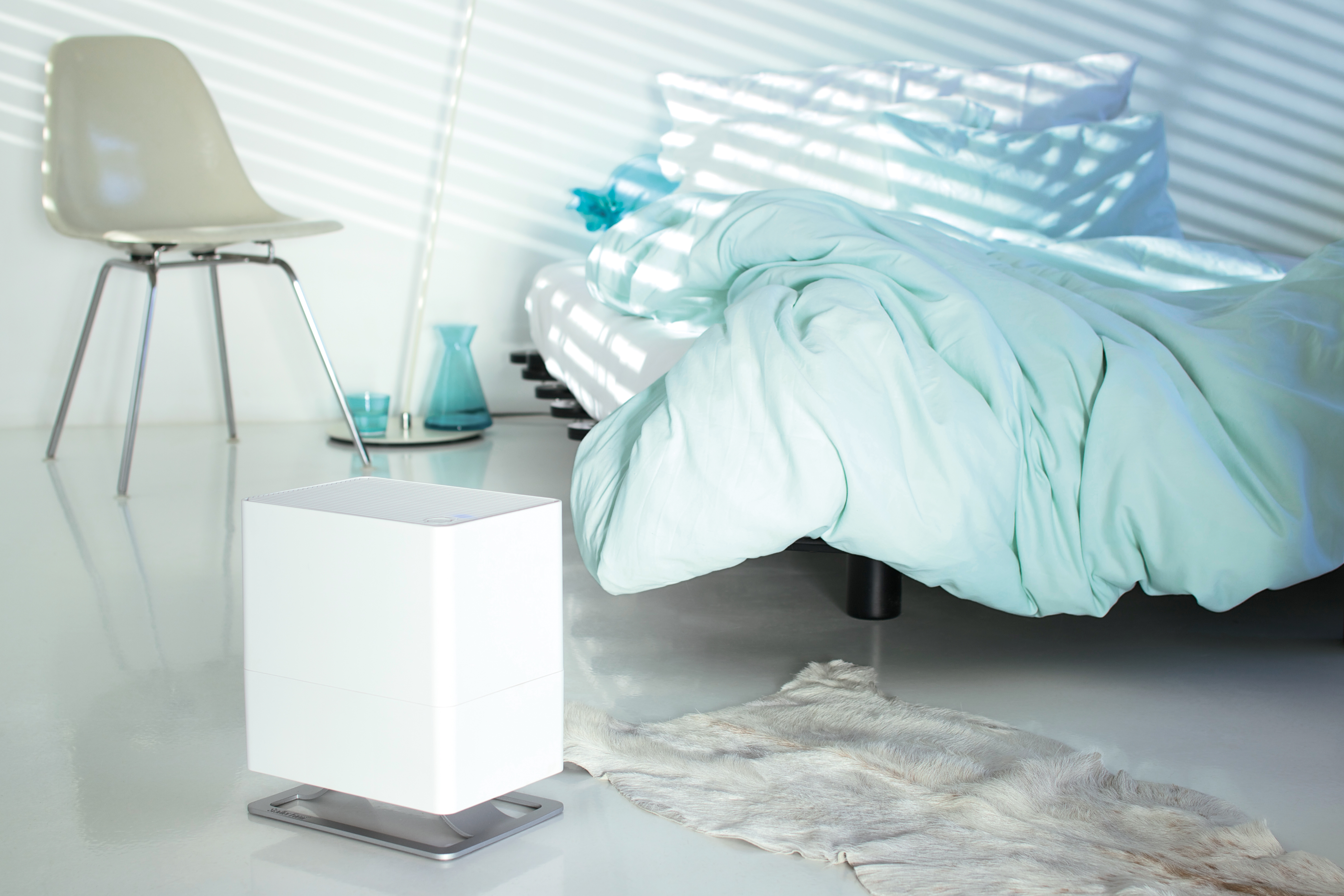 Ein weißer Luftbefeuchter auf einem Ständer in einem Schlafzimmer. Ein Bett mit blauen Decken ist im Hintergrund.