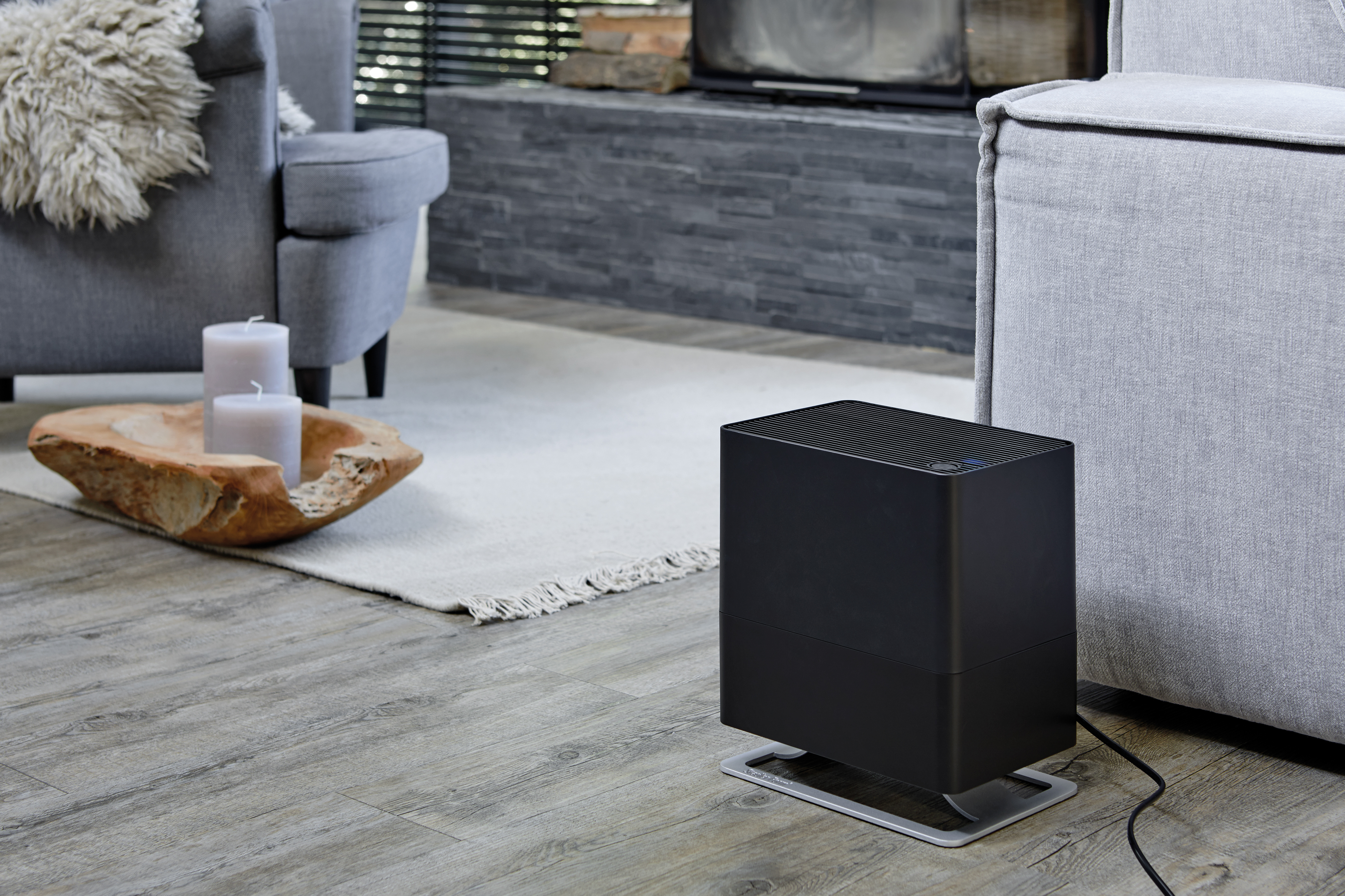 Schwarzer Luftreiniger auf dem Boden, graues Sofa, Holzboden, weißer Teppich und zwei Kerzen in einer Holzschale.