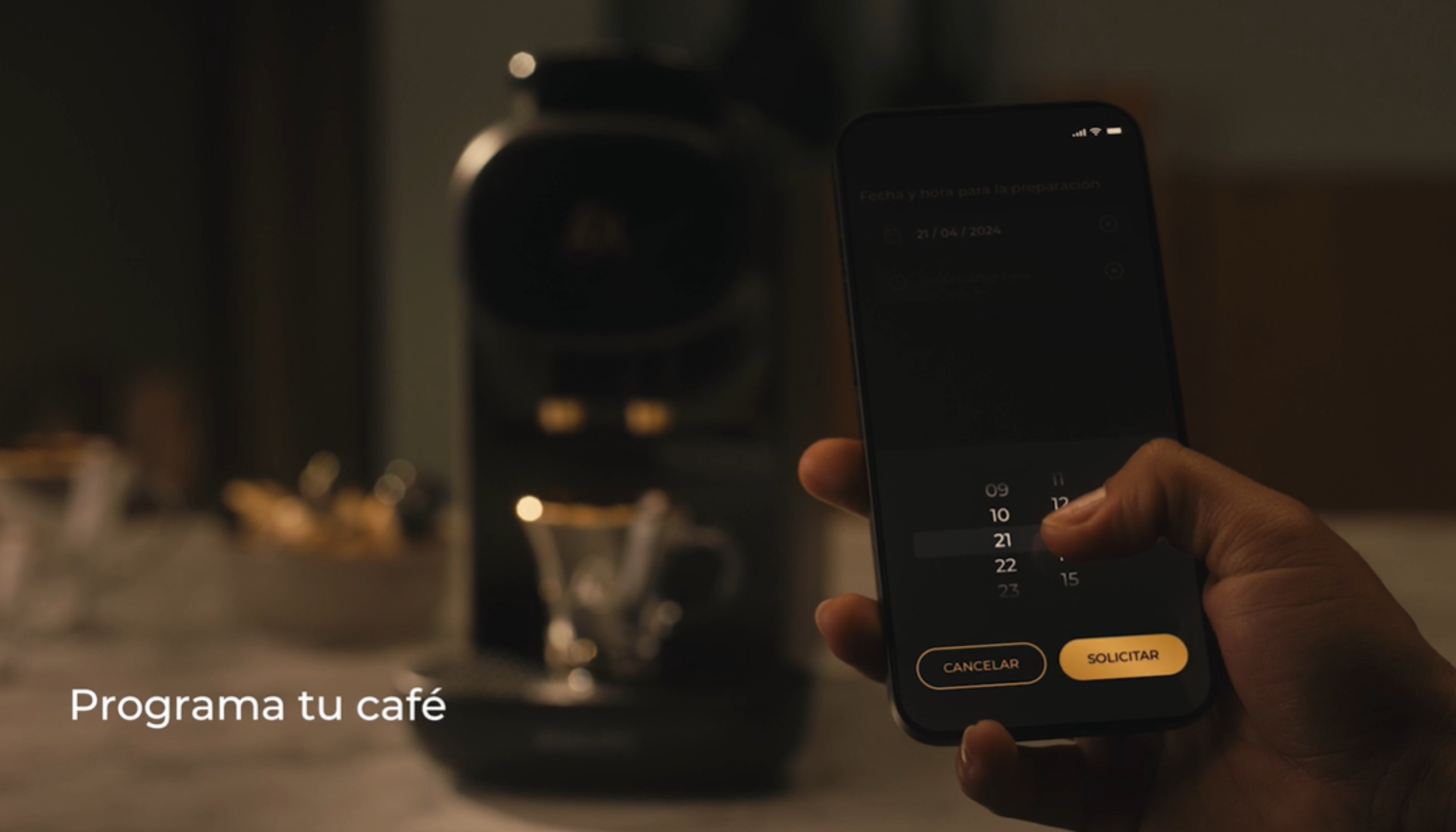 Persona usando teléfono para programar café. Cafetera y taza en el fondo.