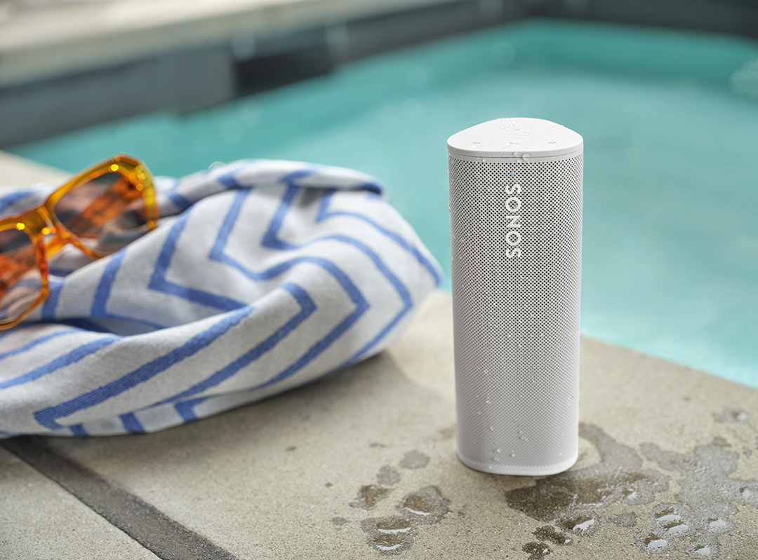 Fehér Sonos hangszóró a medence mellett. Napszemüveg és egy kék-fehér csíkos törölköző a közelben.