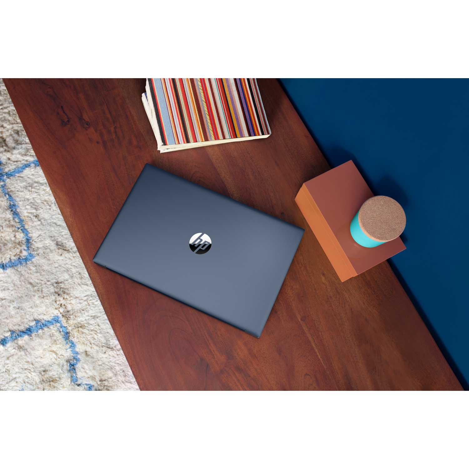 Een donkerblauwe HP-laptop ligt op een houten tafel, met een kleurrijk kunstboek en een klein bakje ernaast.