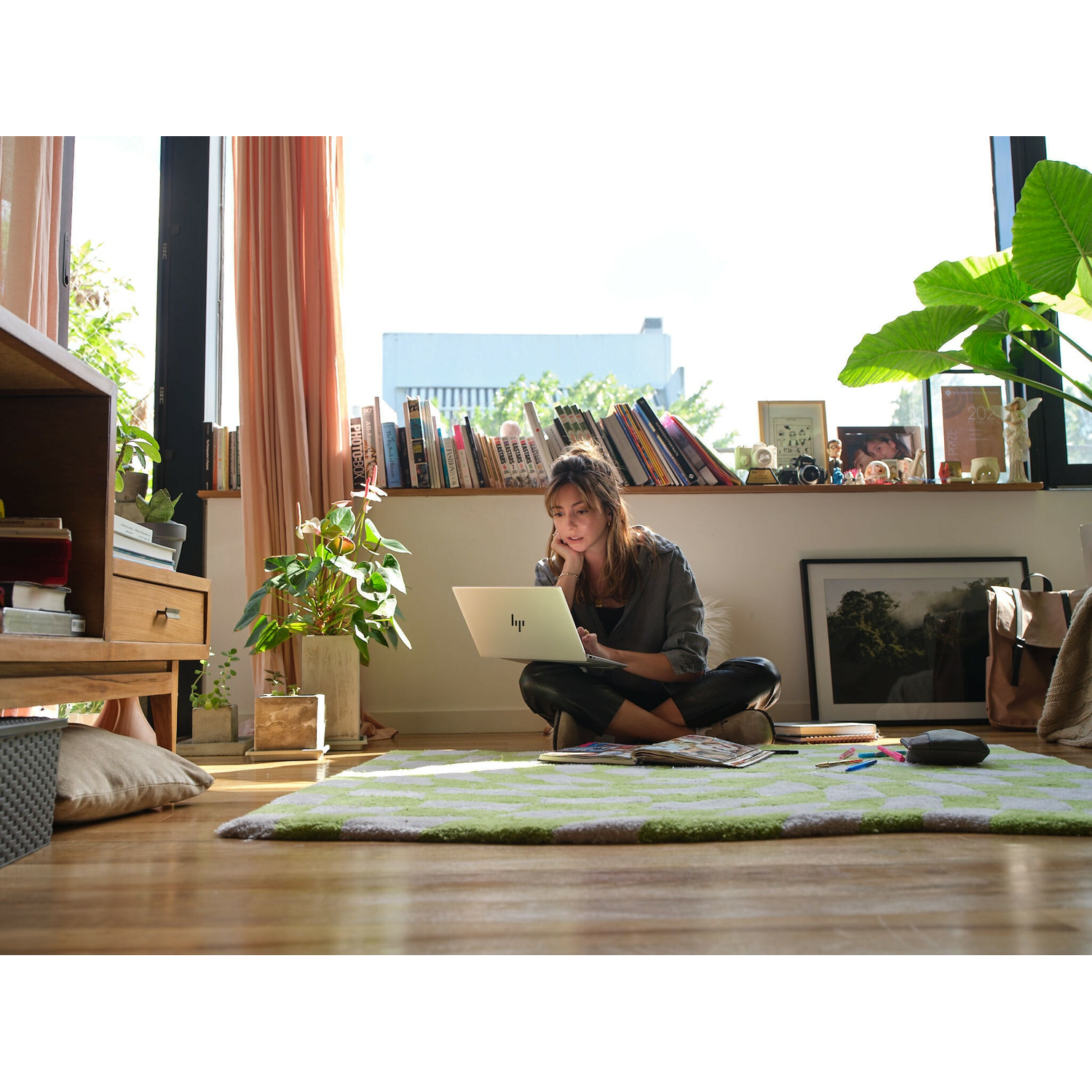 Vrouw zittend op een groen tapijt, werkend op een laptop bij een raam met planten.