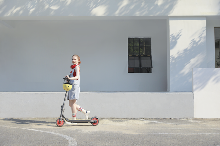 Dziewczyna na hulajnodze, w kasku i słuchawkach, obok białego budynku z oknem.