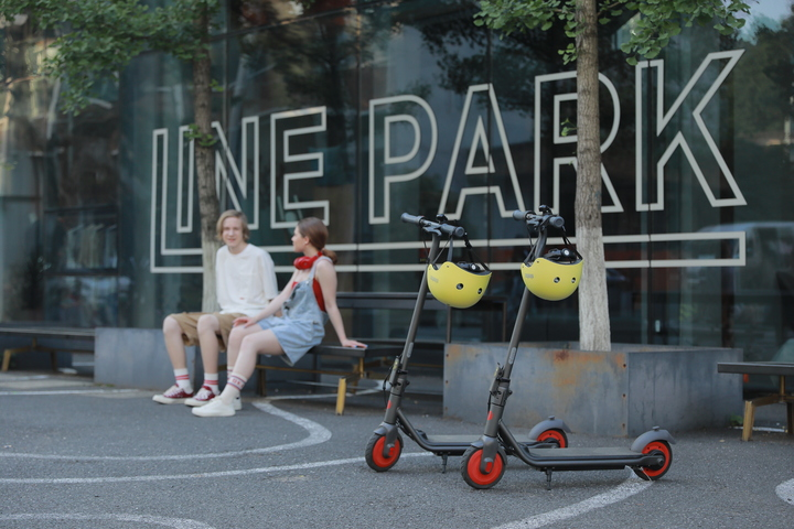 Dwa skutery z żółtymi kaskami zaparkowane przed budynkiem. Ludzie siedzą w tle.