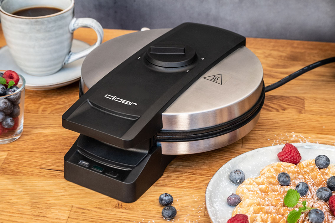 Waffeleisen mit Waffel, Beeren und einer Tasse Kaffee auf einem Holztisch. Schwarz und Silber.