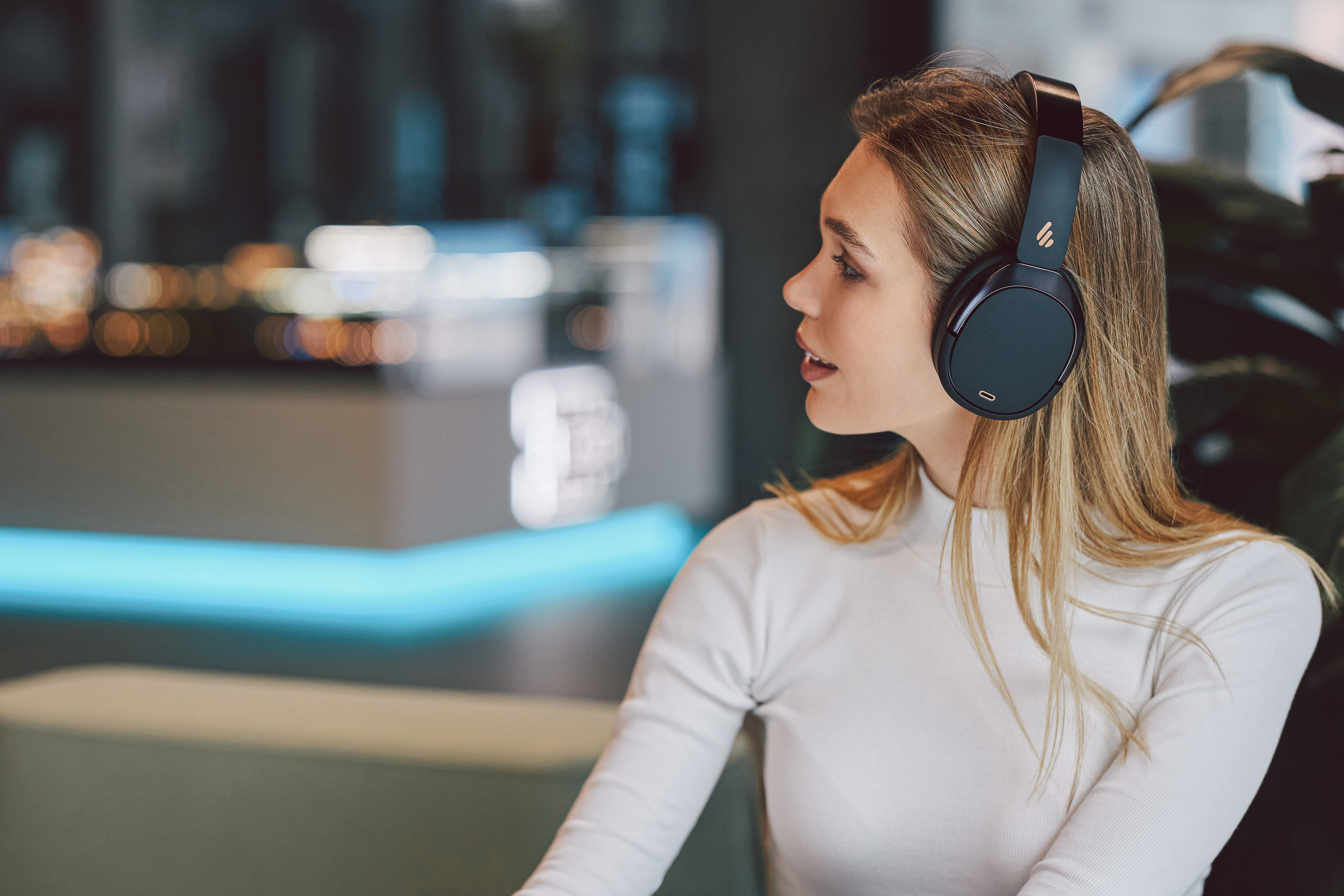 Blonde Frau mit schwarzen Kopfhörern, die zur Seite blickt, mit verschwommenem Café-Hintergrund.