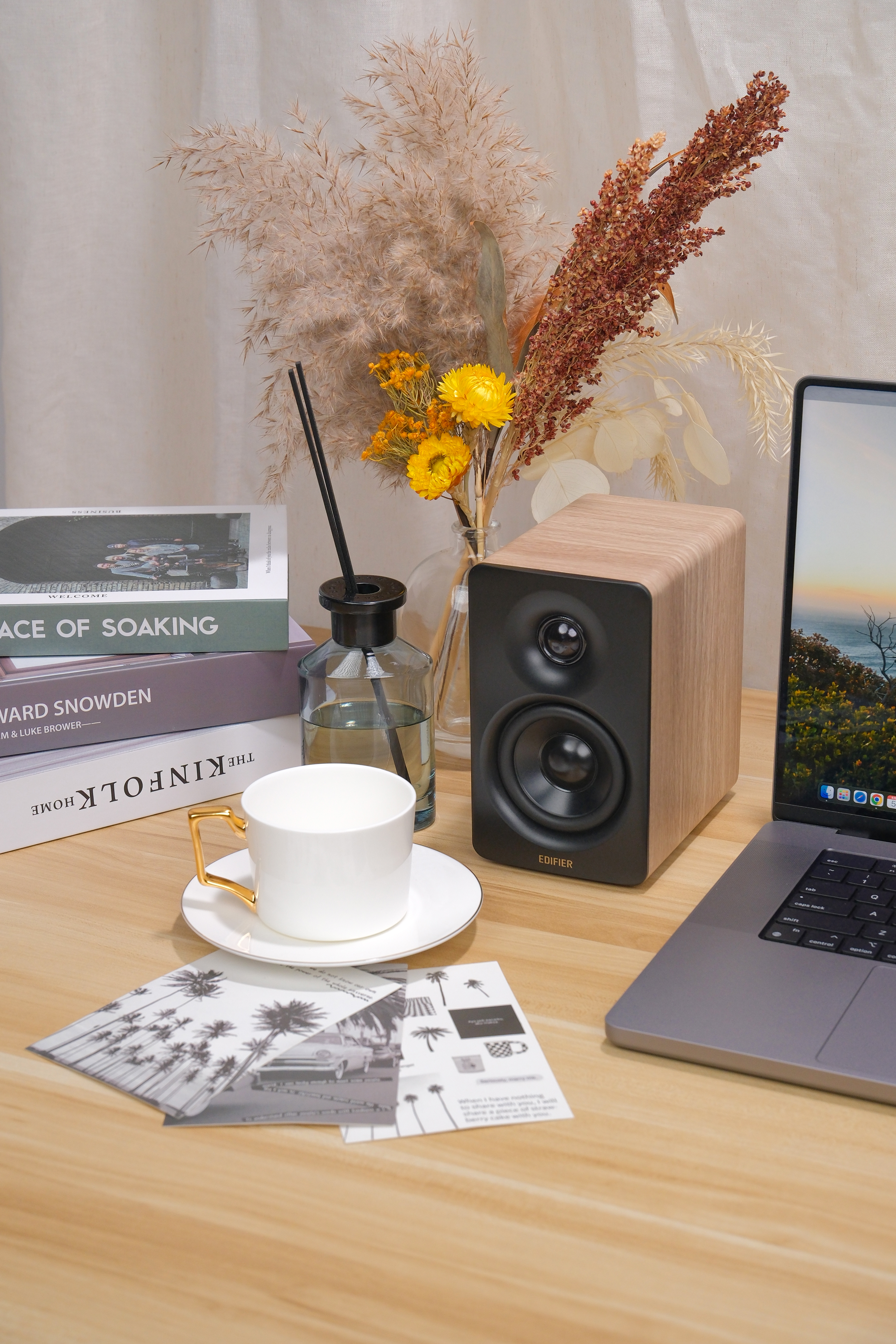 Ein Lautsprecher mit Holzoptik, Bücher, eine Kaffeetasse und ein Laptop auf einem Schreibtisch.