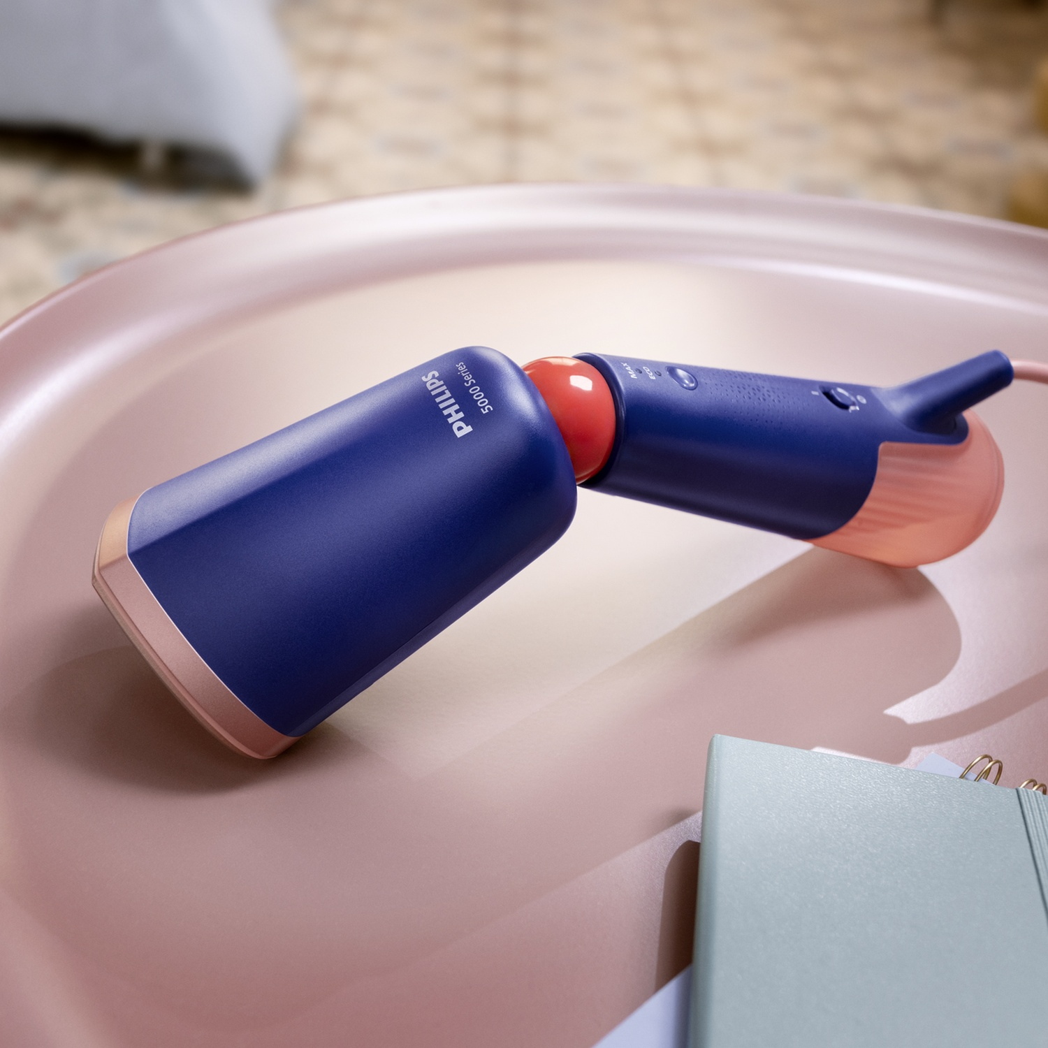 Un vaporizzatore portatile Philips blu e rosso su una superficie rosa, con un taccuino sullo sfondo.