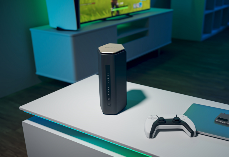 Ein schwarzer und grauer Router auf einem weißen Tisch mit einem Gamecontroller. Im Hintergrund ist ein Fernseher zu sehen.