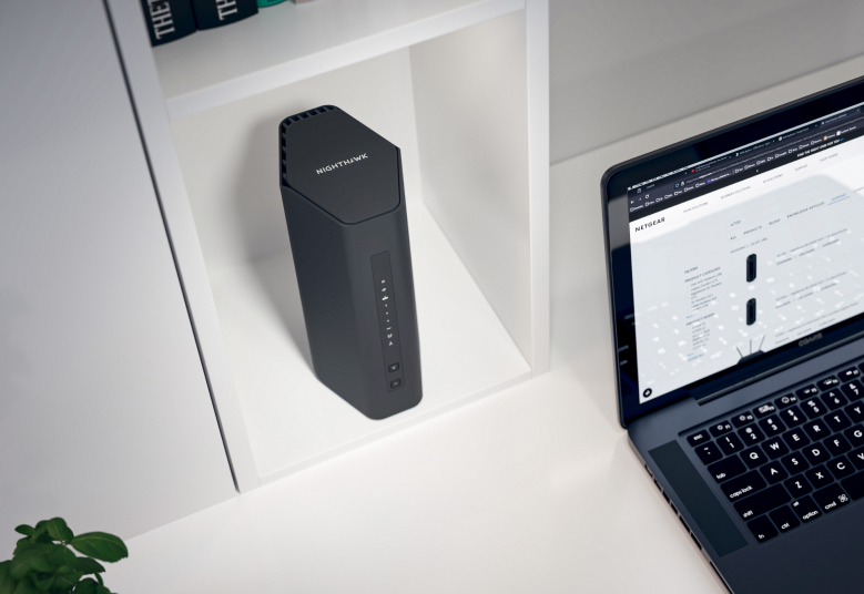 Ein schwarzer Nighthawk-Router auf einem weißen Regal neben einem Laptop, der eine Website anzeigt.