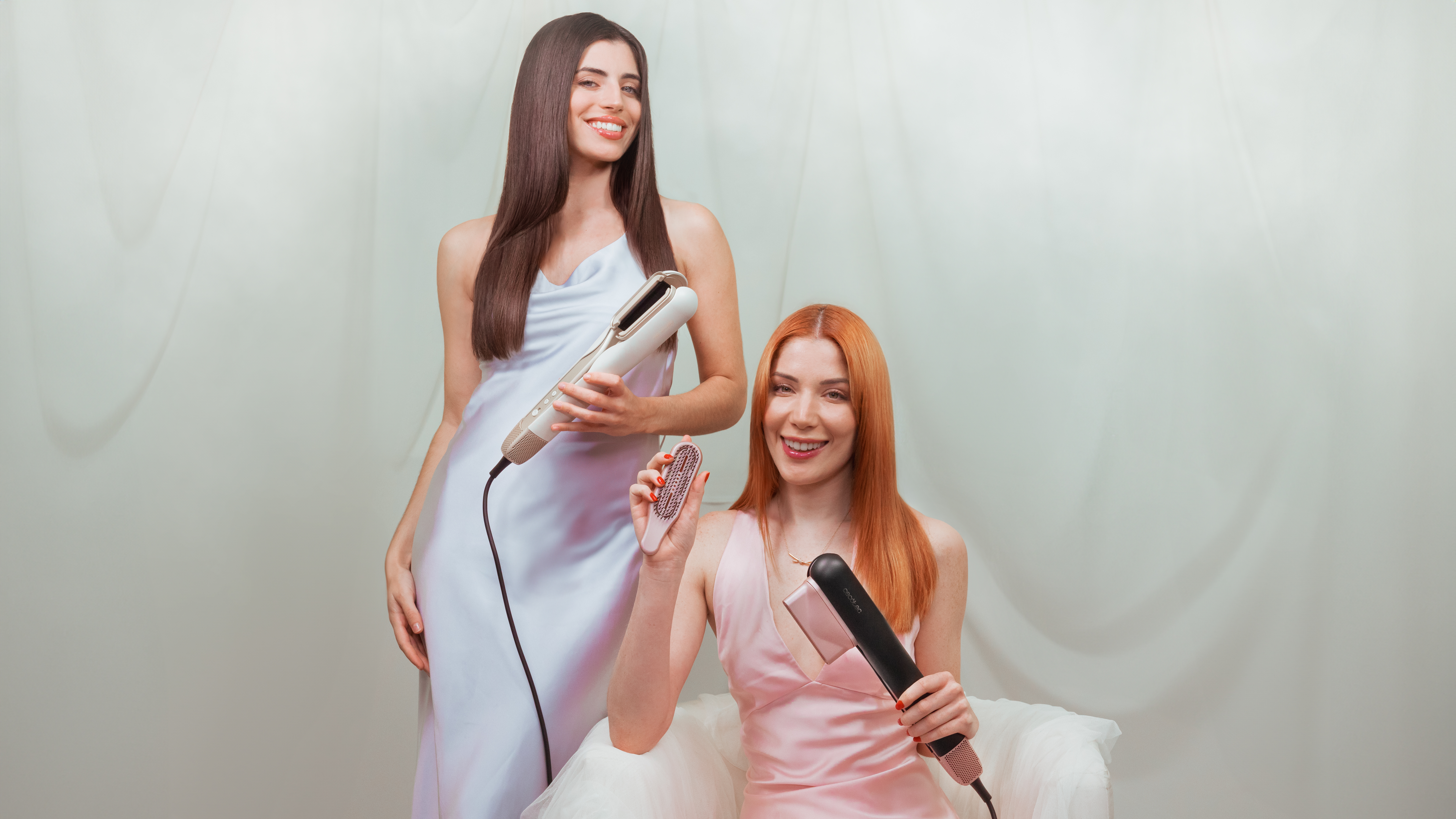 Dos mujeres sonriendo, sosteniendo herramientas para peinar el cabello. Una con vestido blanco, otra con vestido rosa.