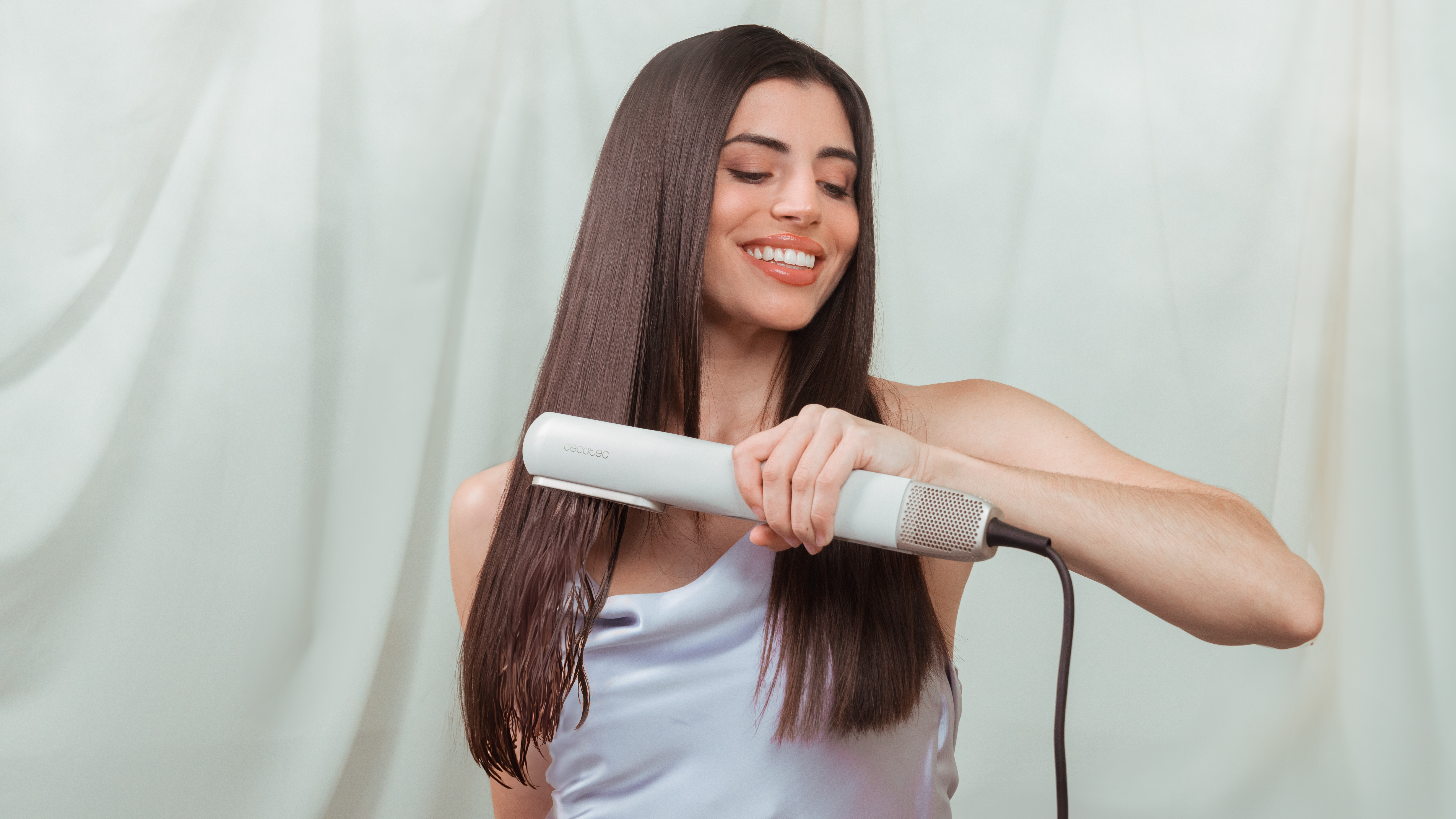 Una mujer con cabello largo usa una plancha de pelo blanca. Está sonriendo y lleva una blusa azul claro.