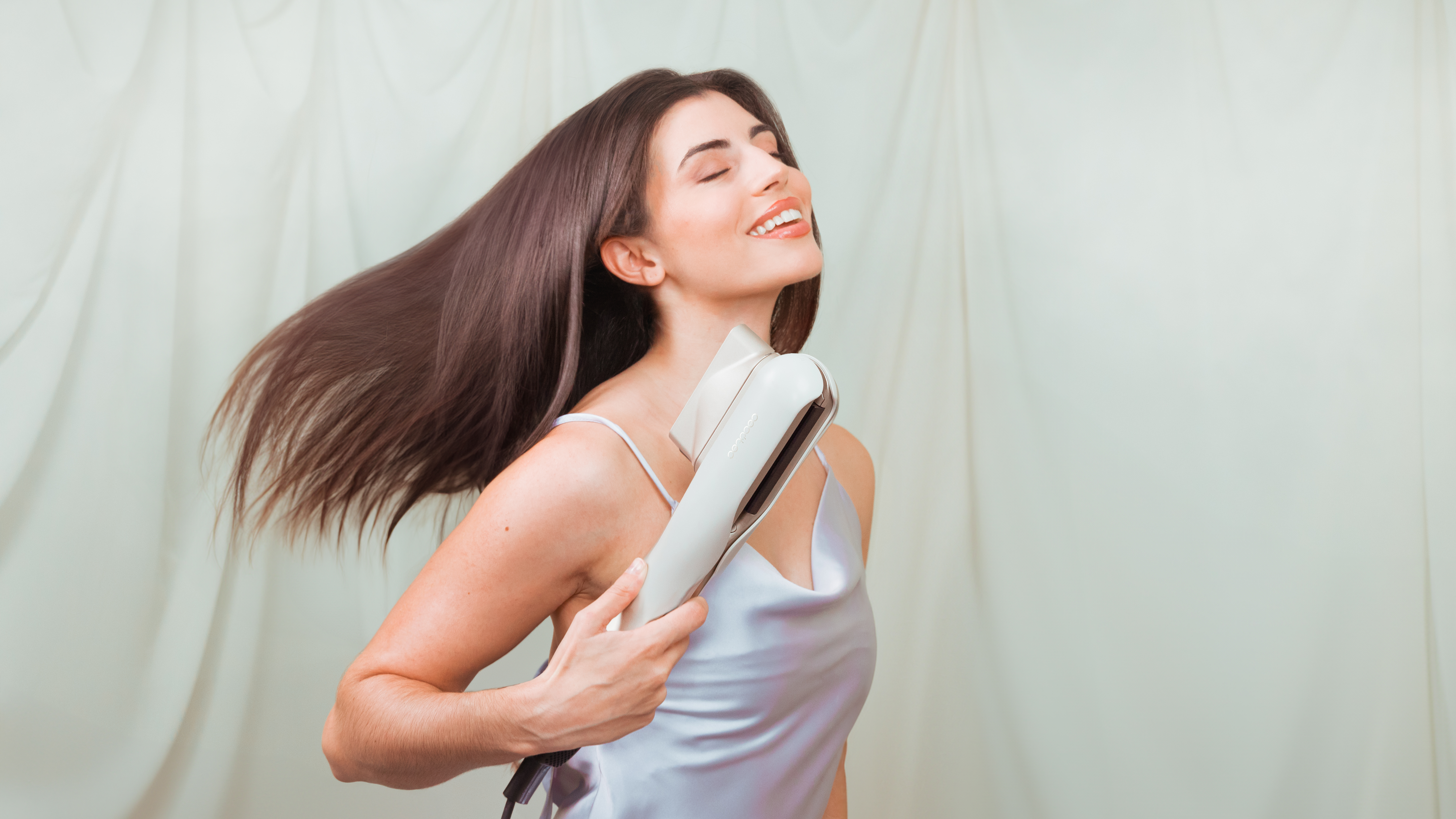 Mujer con pelo largo usando una plancha de pelo, sonriendo y mirando hacia arriba. Cortinas cremosas en el fondo.
