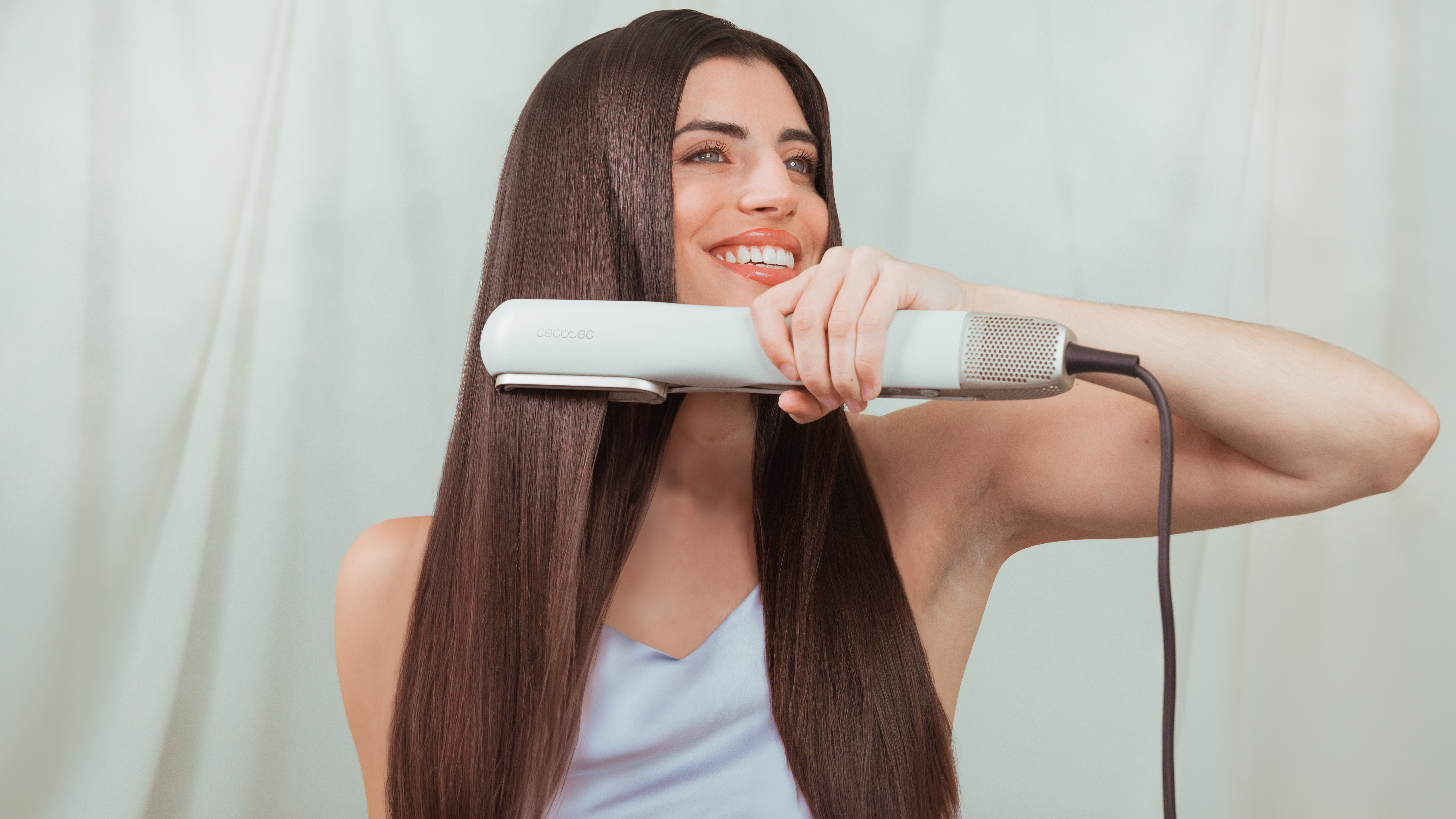 Mujer alisando el cabello con una herramienta de peinado. Sonríe y mira hacia arriba.