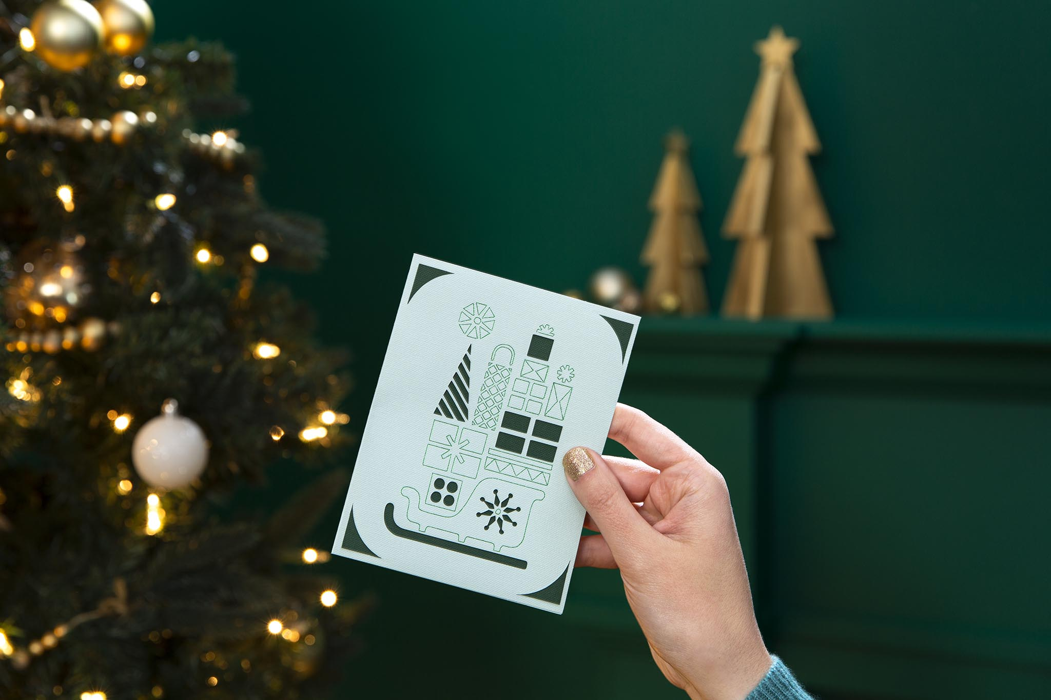 Hand hält Weihnachtskarte mit Geschenken auf einem Schlitten, Weihnachtsbaum und Hintergrund.