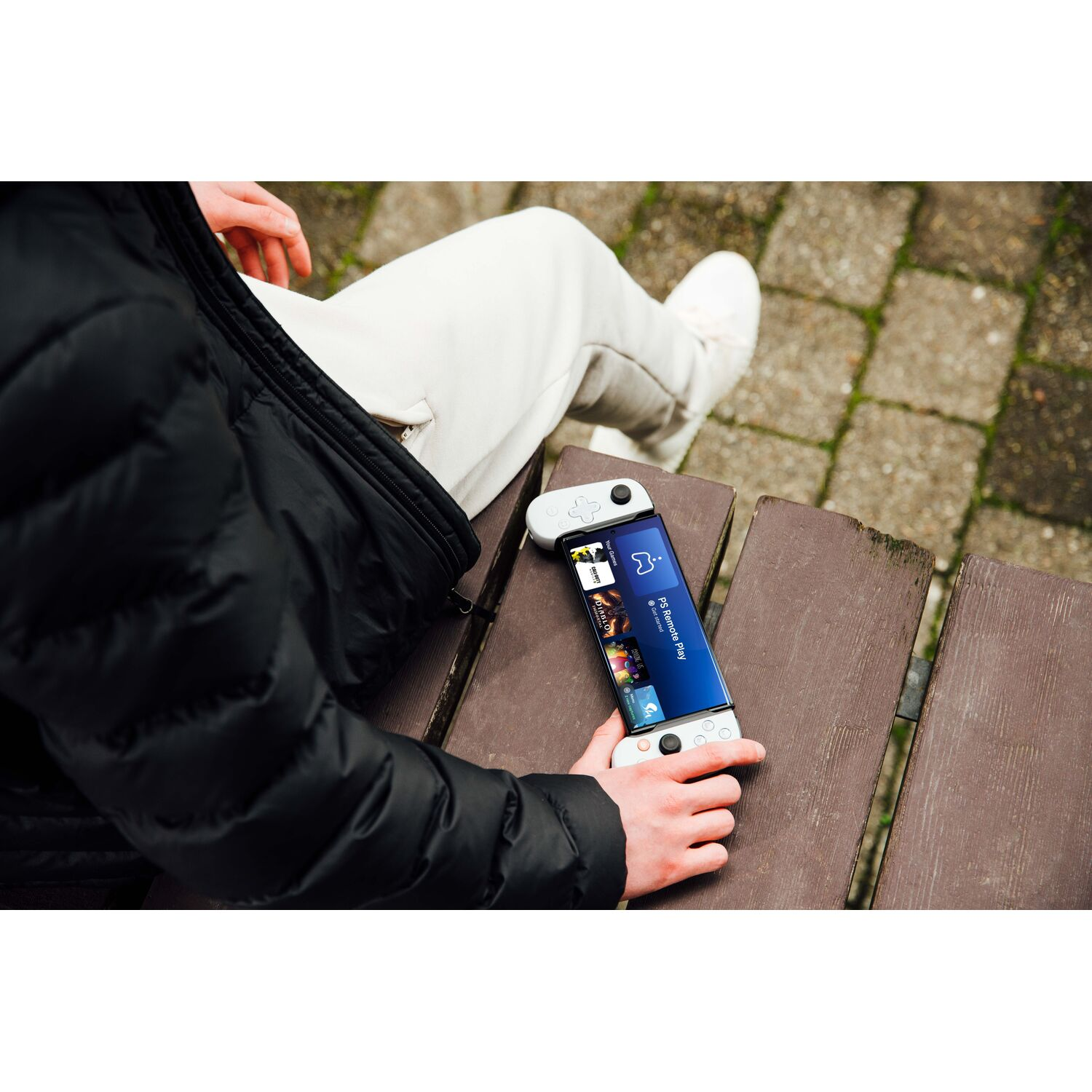 Person holds a phone with attached game controllers, sitting on a wooden bench. Background is blurred.