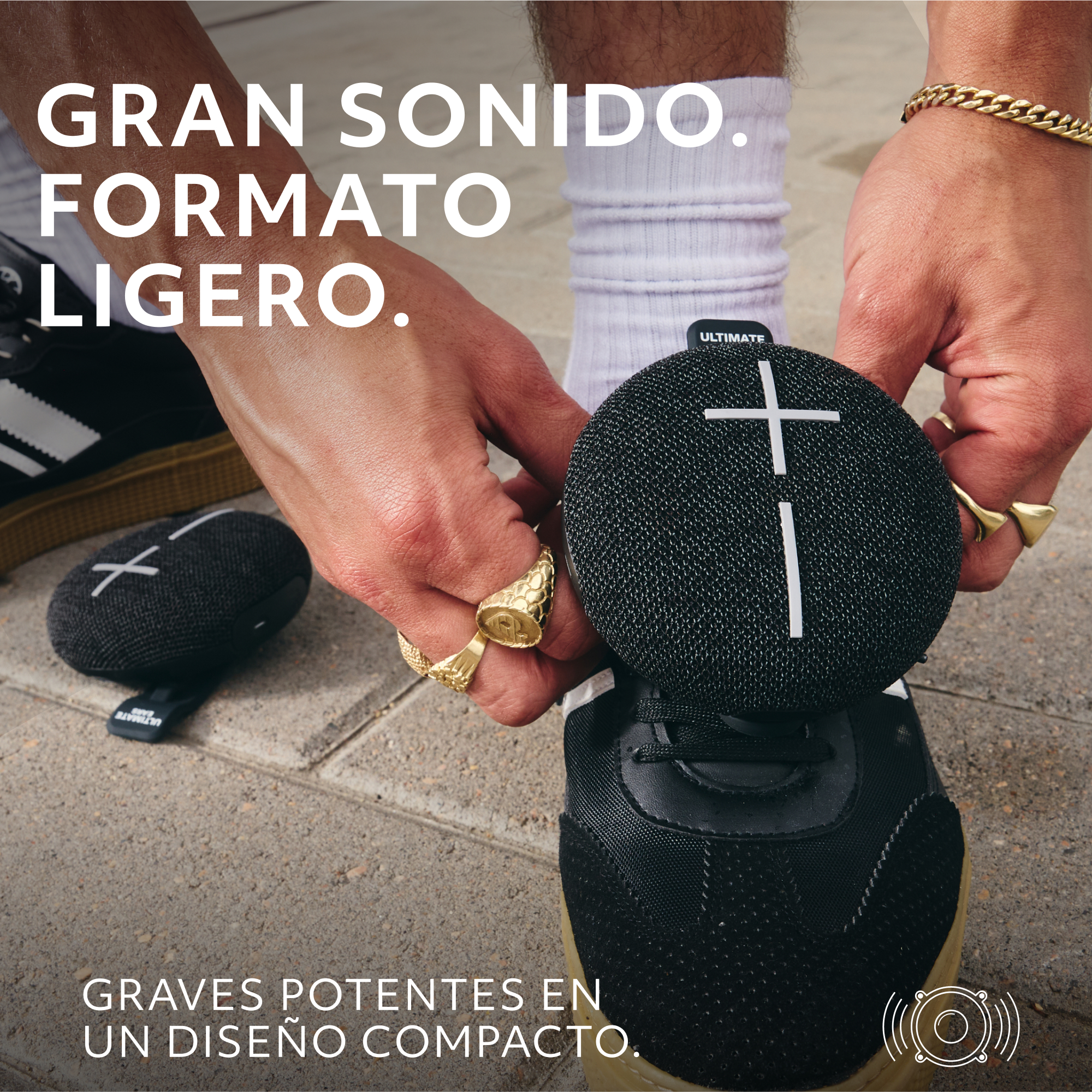 Un altavoz negro sobre un zapato. Manos sosteniéndolo. Texto en español: Sonido grande, forma ligera, bajos potentes en diseño compacto.