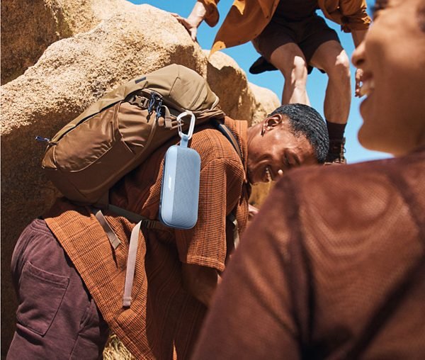 Grupa ludzi na wędrówce, jeden niesie niebieski głośnik, wspinający się po skalistym terenie.