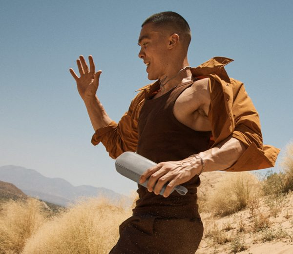 Mężczyzna na pustyni, trzymający szary głośnik, podnosi rękę. Brązowa koszula, beżowe spodnie, uśmiech.