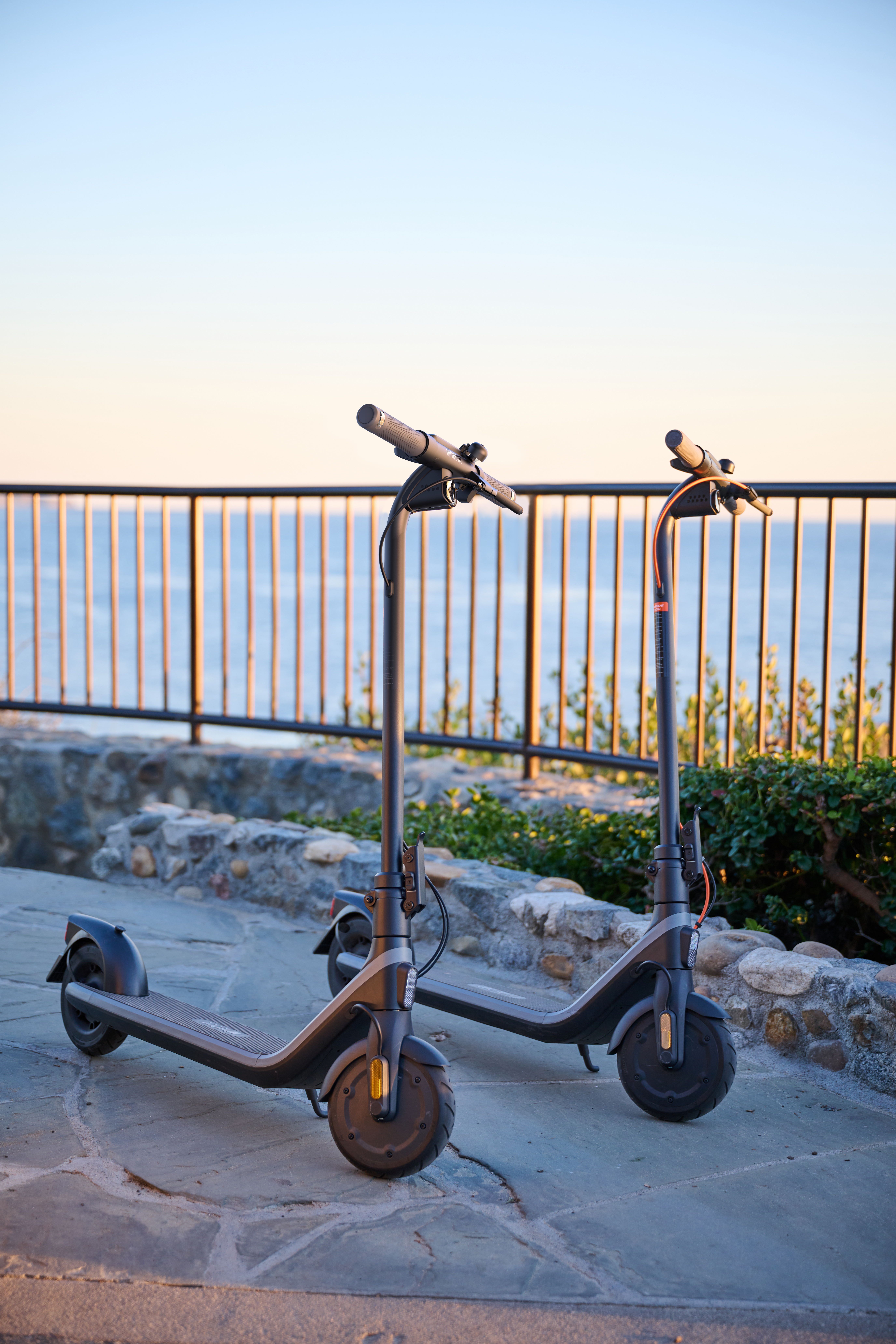 Dwa szare skutery elektryczne na kamiennej ścieżce, z metalowym ogrodzeniem i morzem w tle.