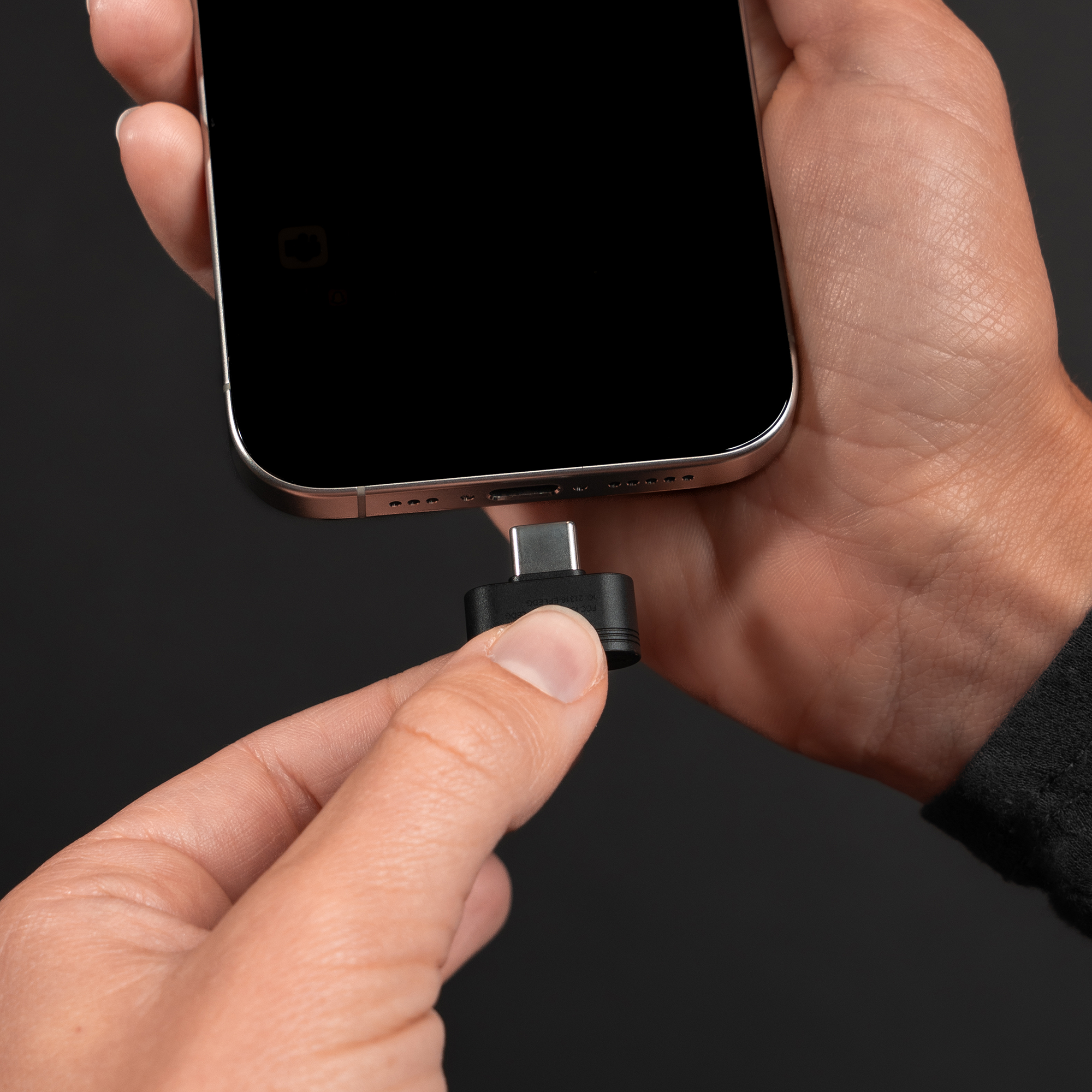 Egy kéz egy USB-C adaptert tart, ami egy telefonhoz csatlakozik, fekete háttér előtt.