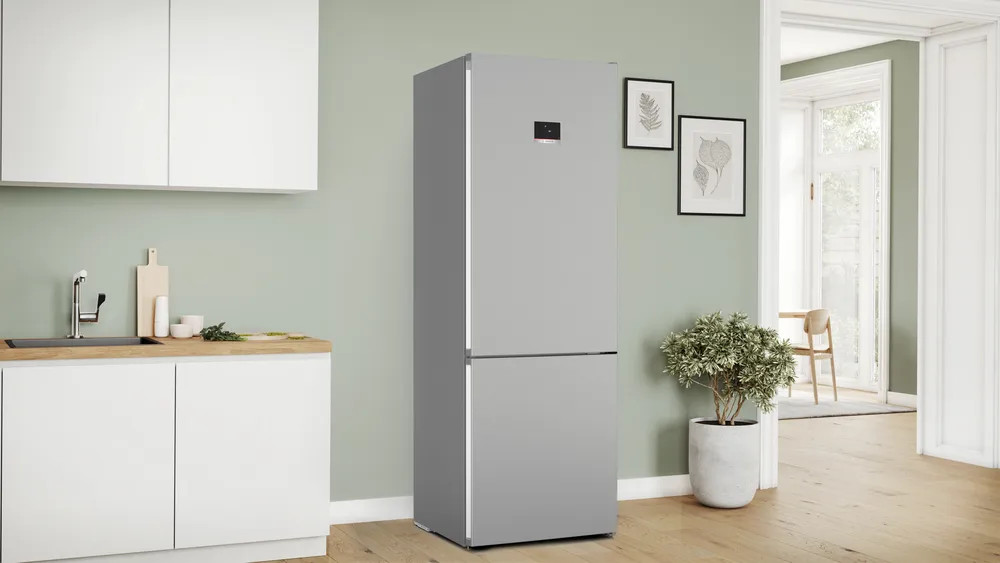 Srebrna lodówka na tle jasnozielonej ściany w kuchni. Zawiera roślinę i oprawioną sztukę.