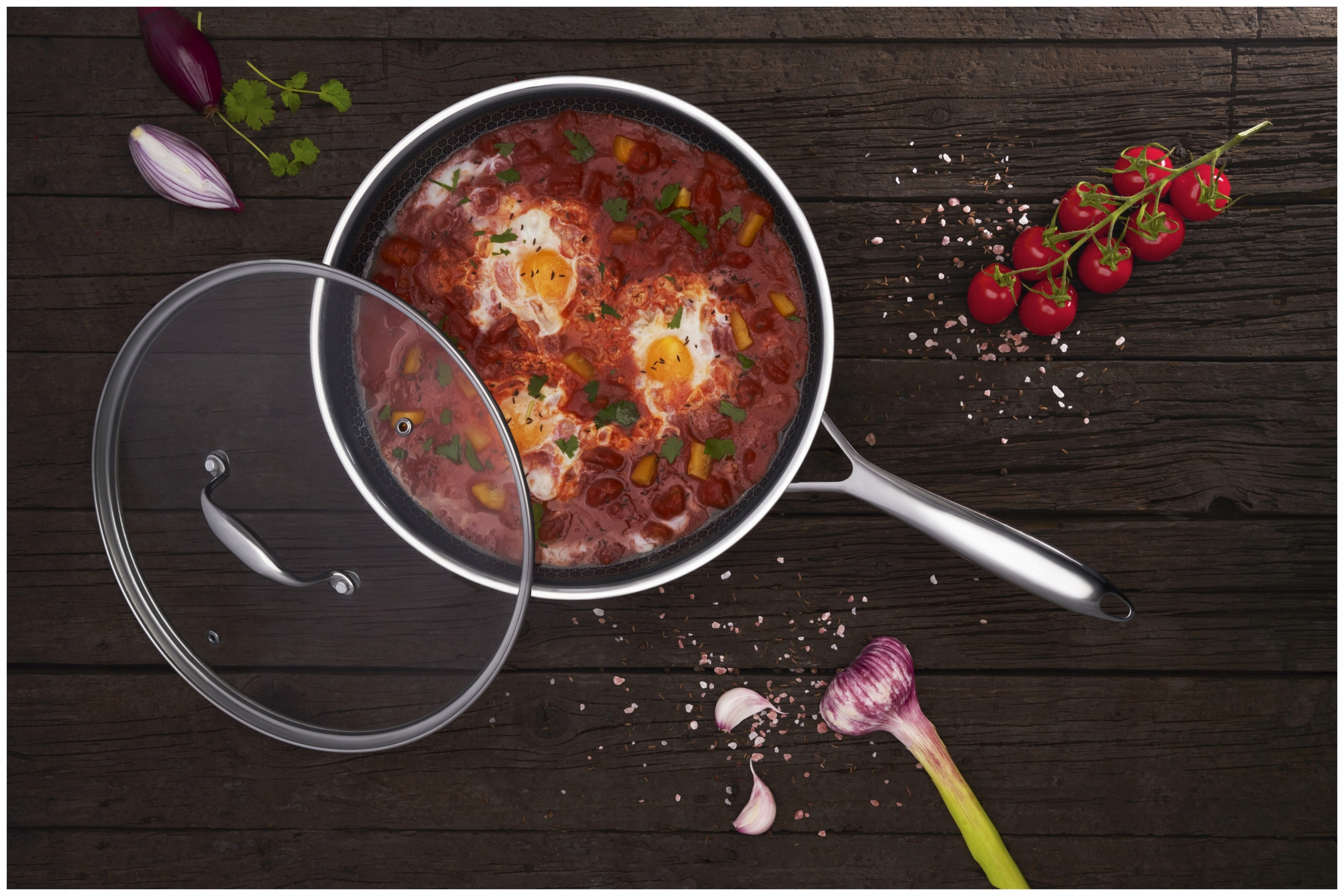 Szakszuka w patelni. Jajka w sosie pomidorowym, z pietruszką. Składniki i pokrywka na drewnianym stole.