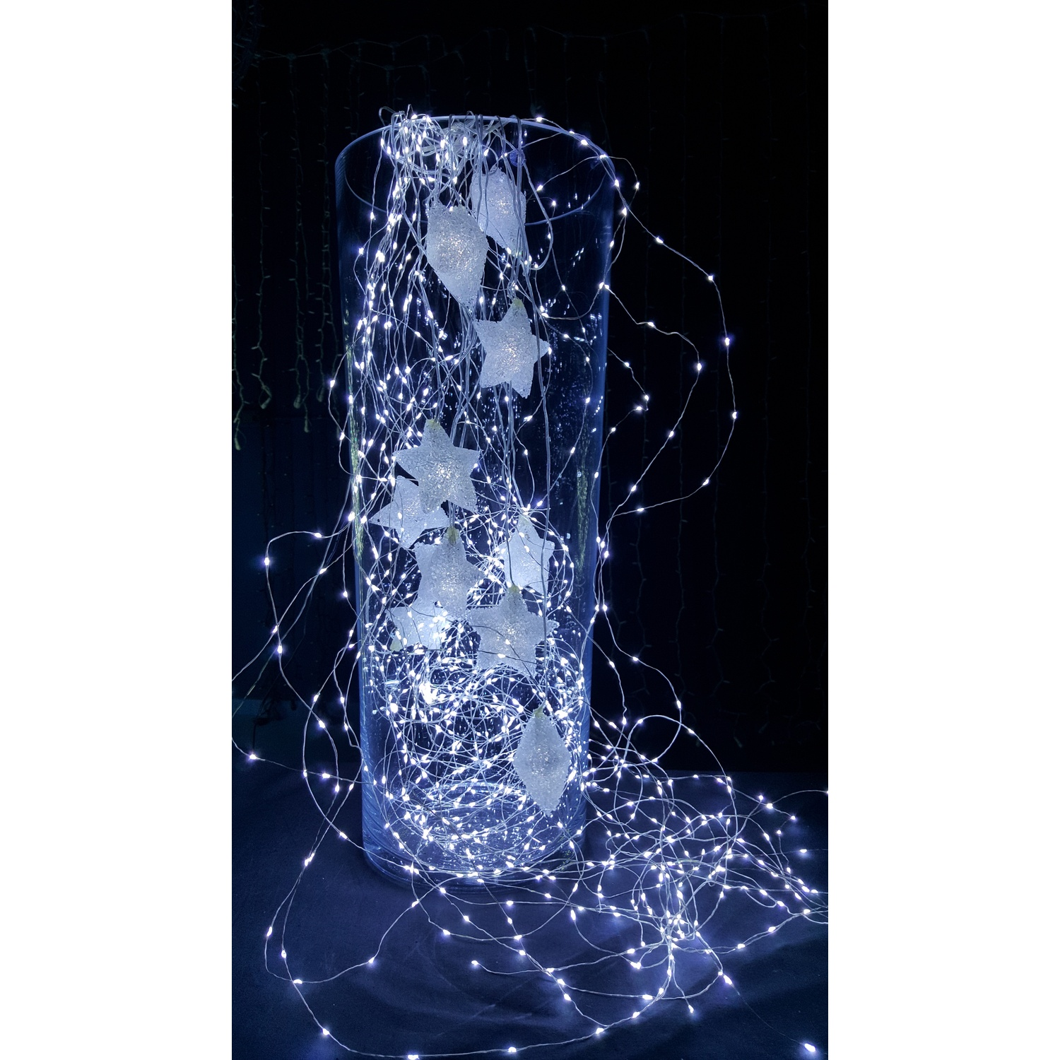 Un vaso di vetro trasparente con luci bianche e ornamenti a forma di stella su sfondo nero.