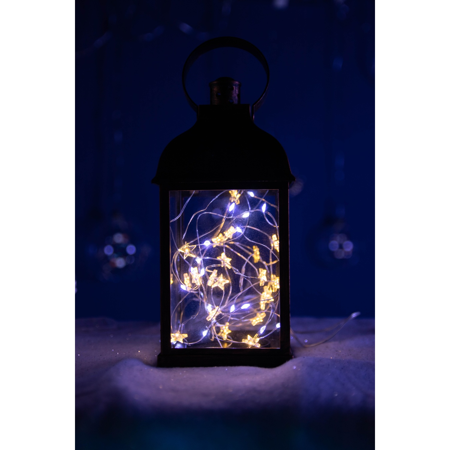 Lanterna nera con luci a stella all'interno. È su una superficie bianca con sfondo blu.