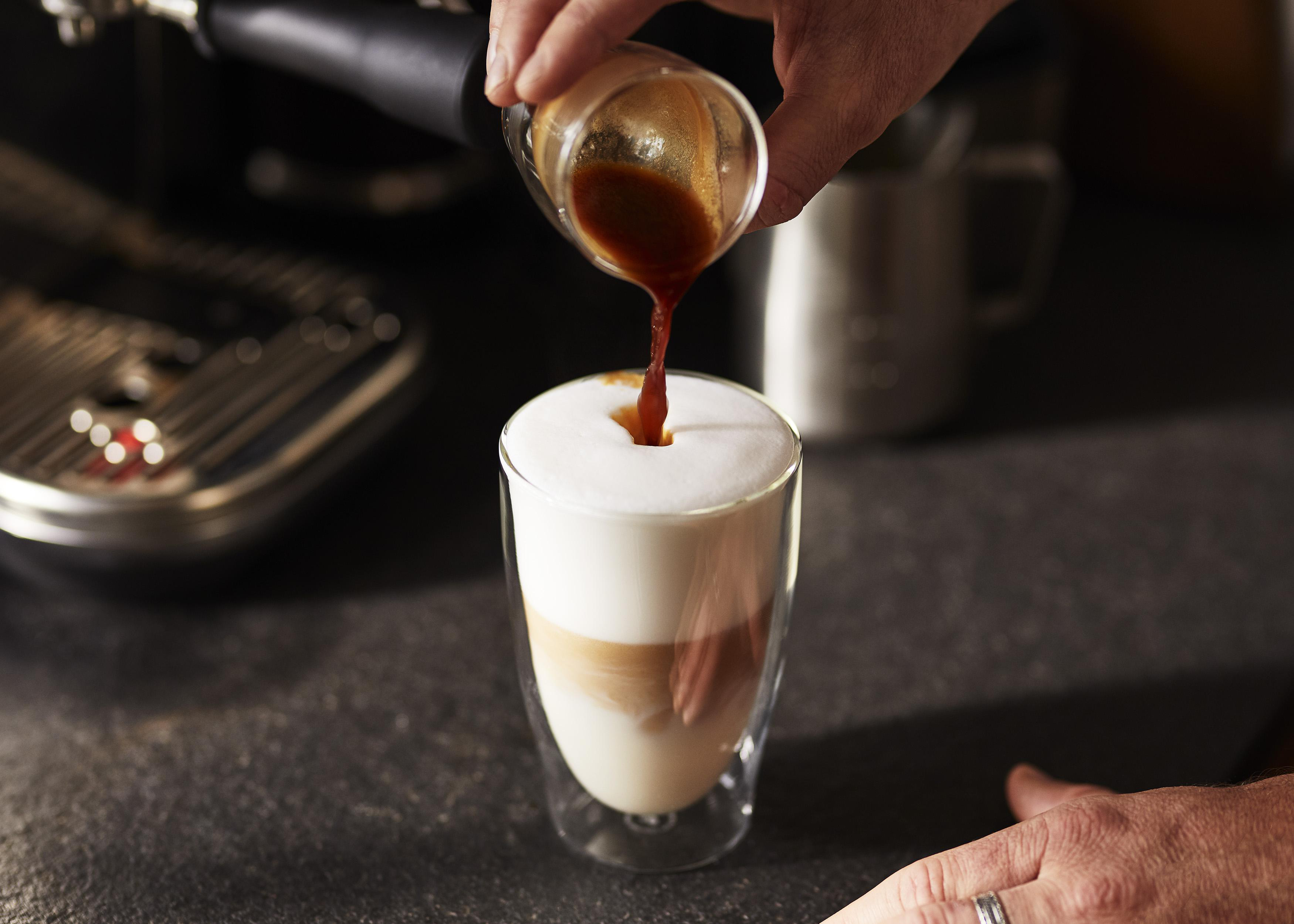 Hand gießt Espresso in ein Glas mit geschichteter Milch und Schaum.