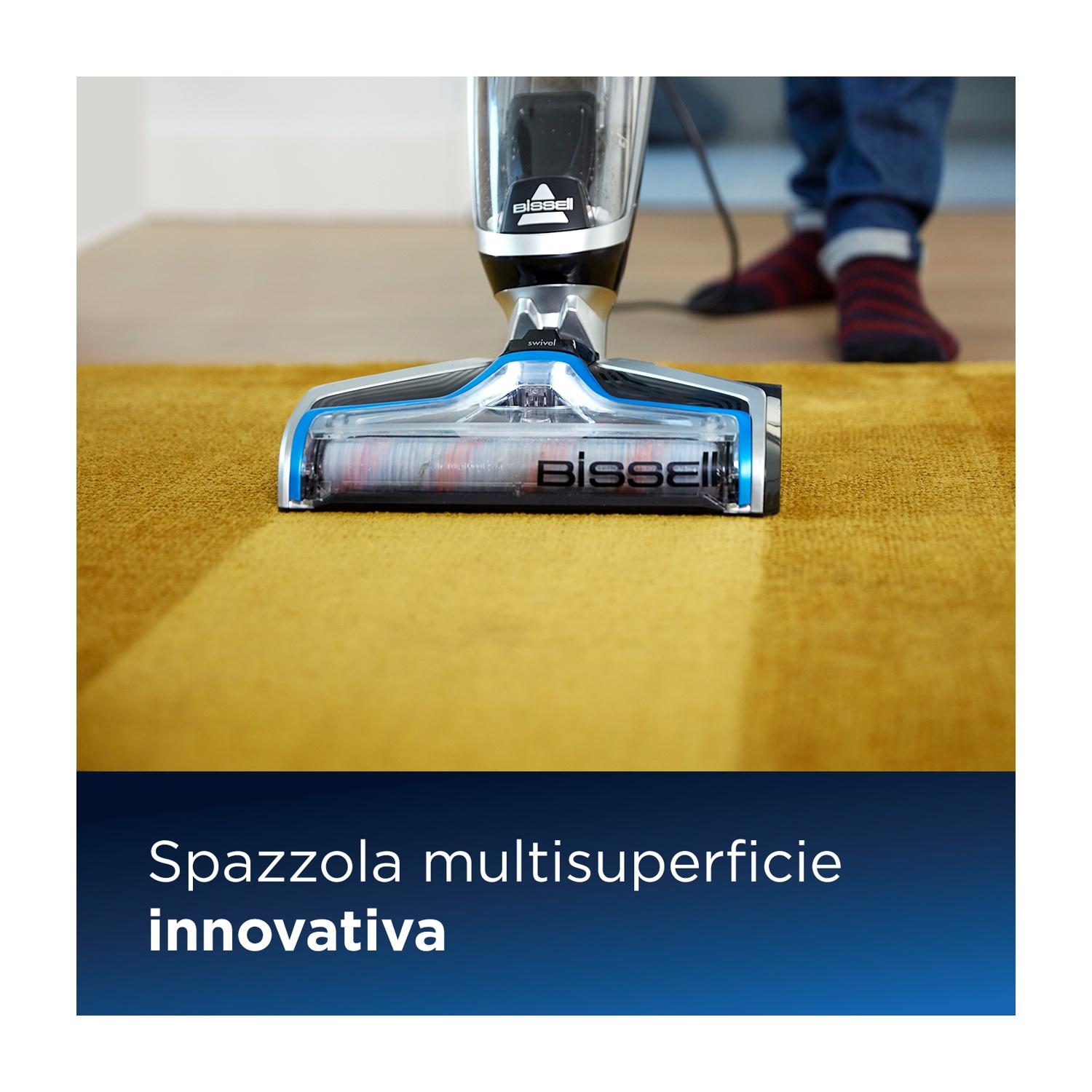 Un dispositivo per la pulizia dei pavimenti pulisce un tappeto giallo; una persona con calzini sullo sfondo.