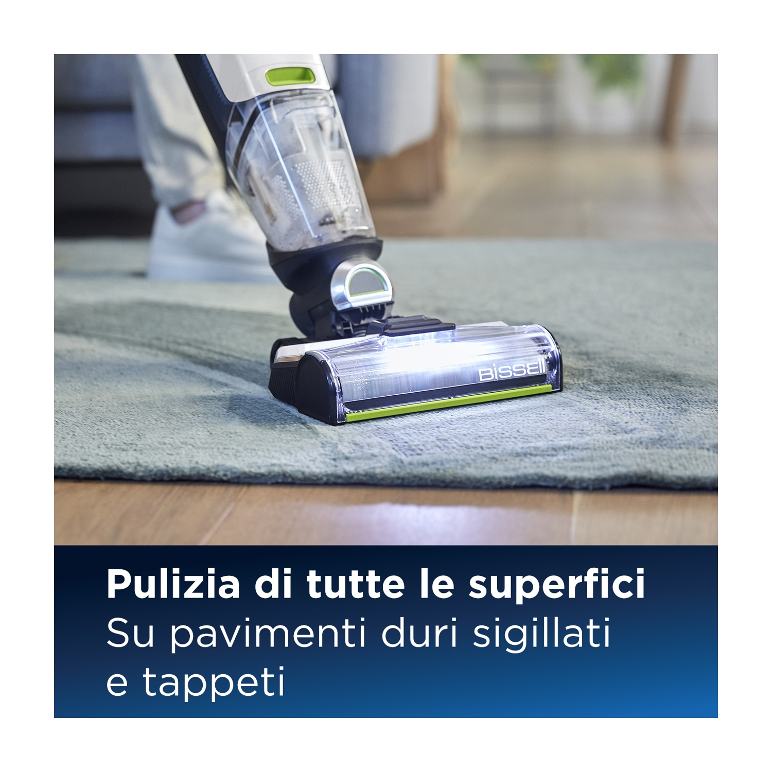 Testa aspirapolvere pulisce tappeto verde, gambe di una persona sullo sfondo.