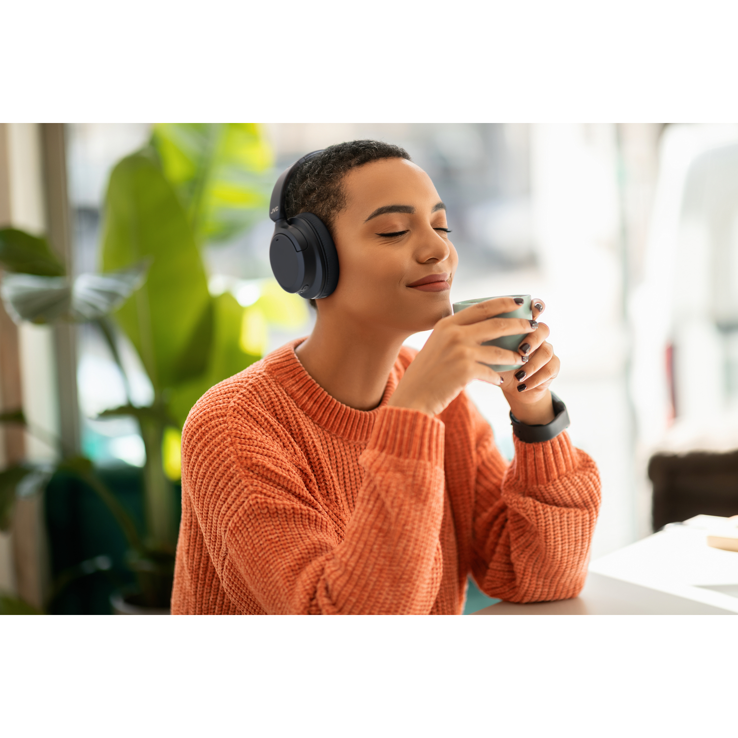 Woman wearing black headphones, holding a mug, in a cafe. Sunlight.