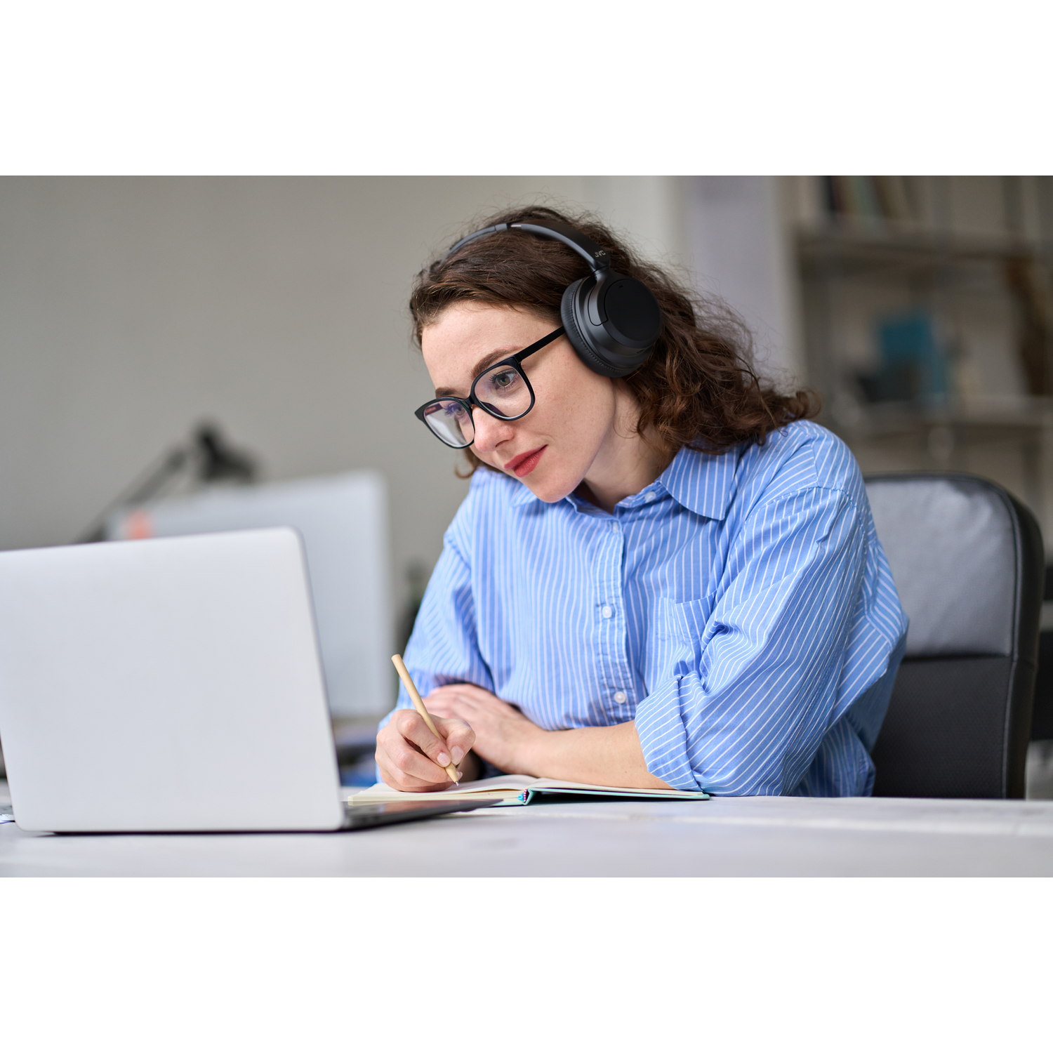 Woman wearing glasses and headphones, writing on a laptop, with a pen and notepad.