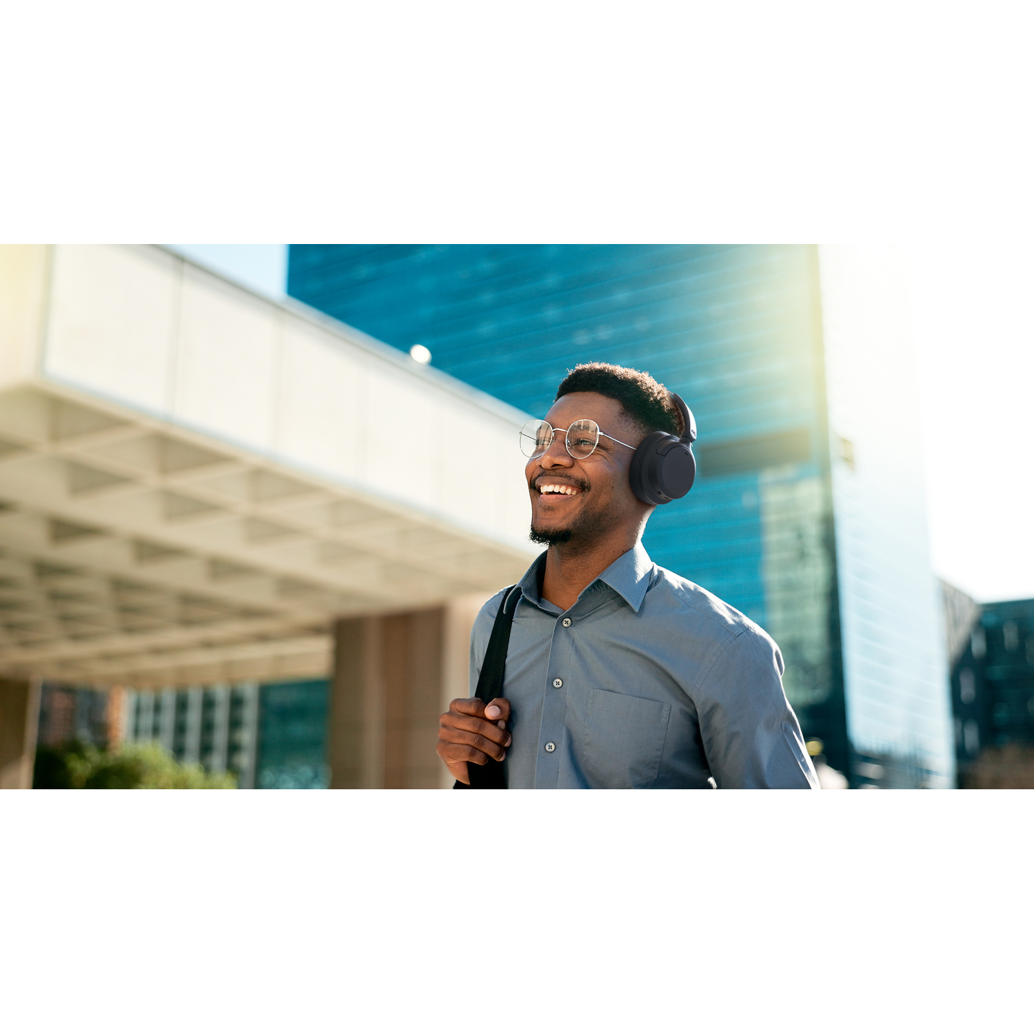 Man wearing headphones smiles outdoors, with buildings in background.