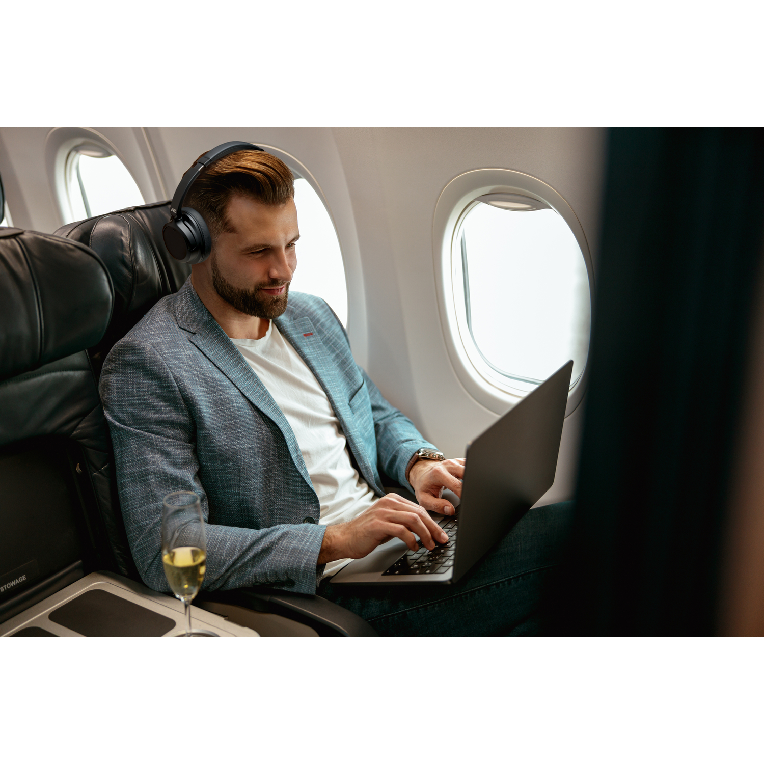 A man in a suit on a plane using a laptop, with a glass of wine on the table.