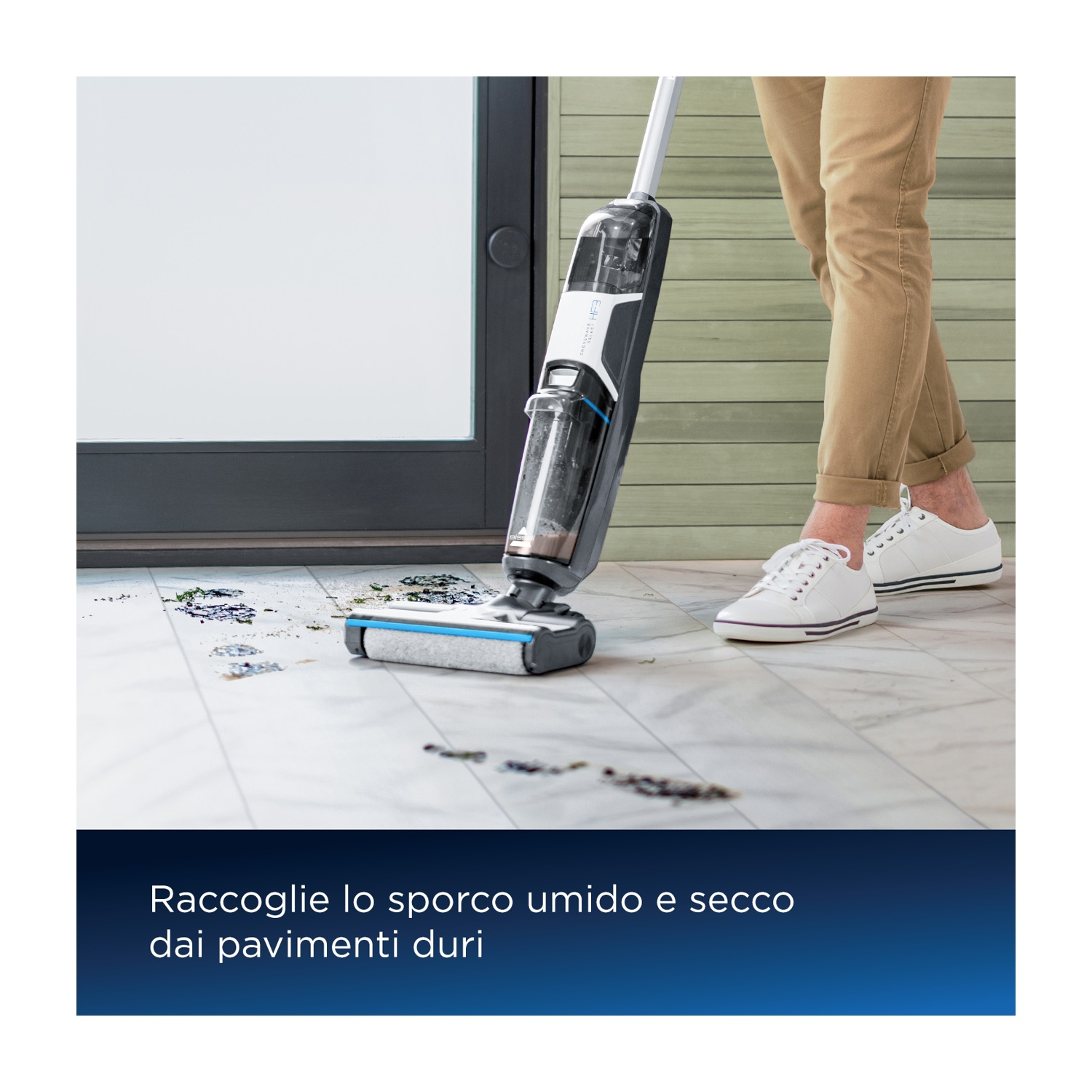 Una persona usa un aspirapolvere su un pavimento piastrellato, pulendo detriti umidi e secchi. Scarpe bianche visibili.
