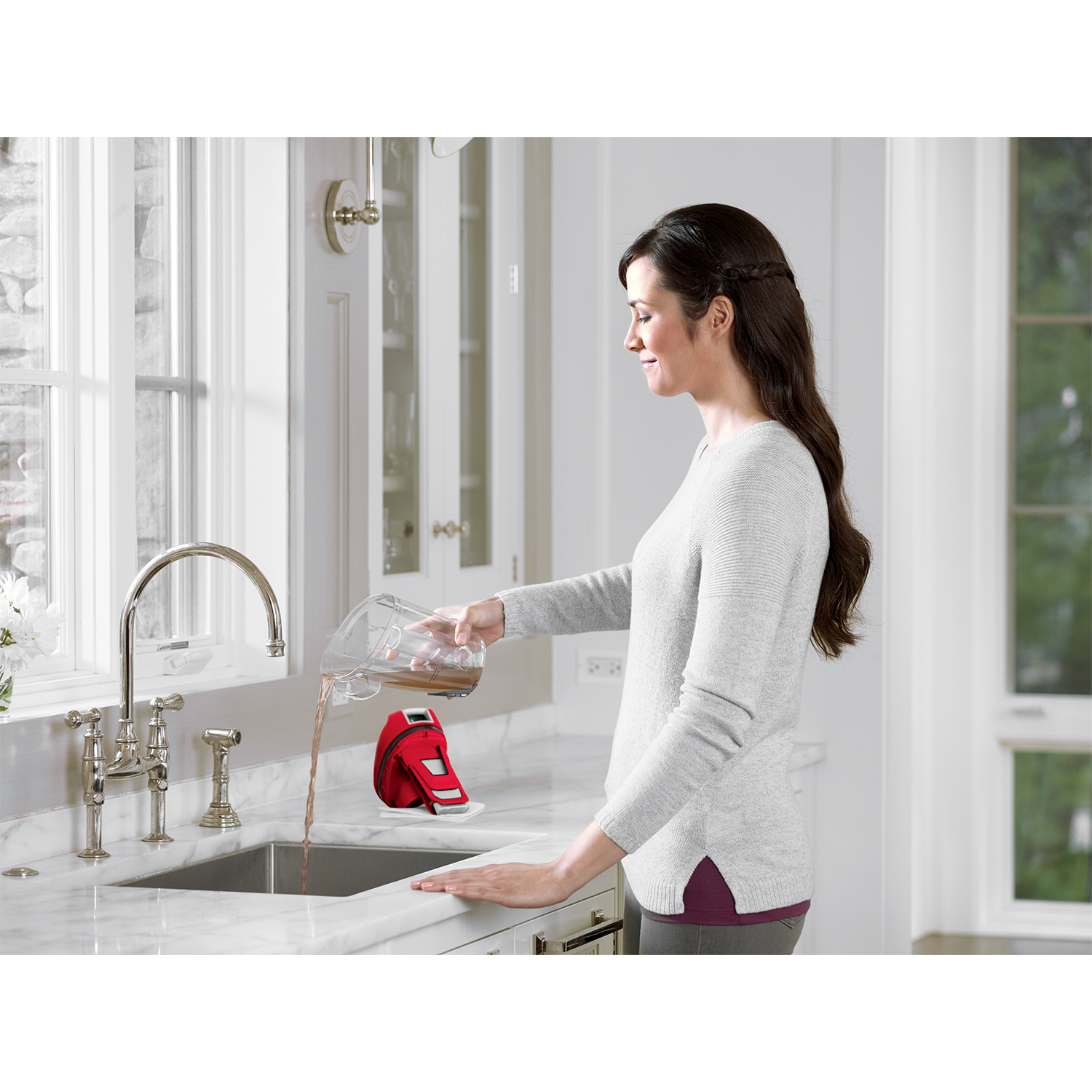 Donna che versa acqua da una brocca in un lavandino, un elettrodomestico rosso è sul bancone accanto al lavandino.