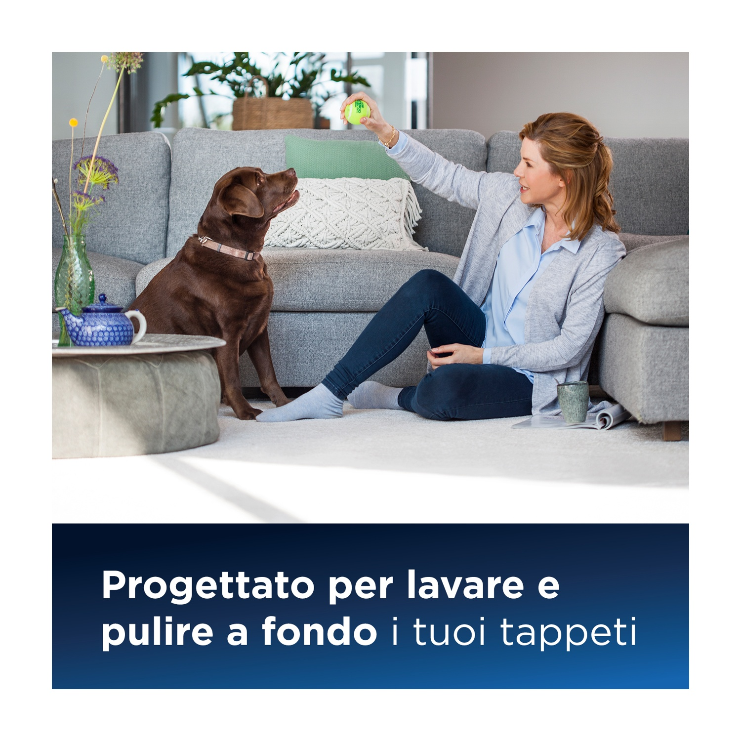 Una donna seduta su un divano gioca con un cane e una palla. La scena è in interni.