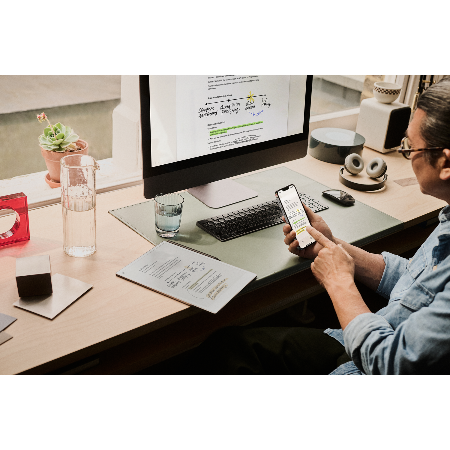 Een persoon werkt aan een bureau. Een computer, telefoon en documenten zijn zichtbaar, de persoon houdt de telefoon vast.