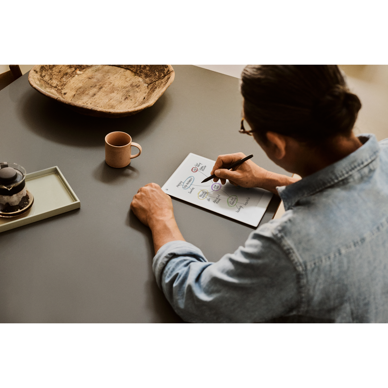 Person sketching on a notepad with a pen, coffee cup and tray on a gray table.