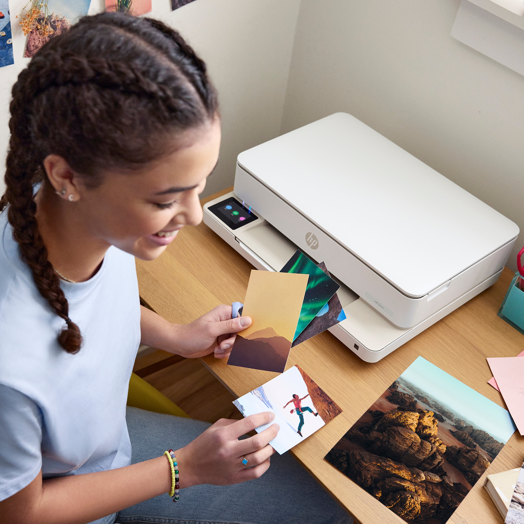 Eine junge Frau lächelt und hält Fotos vor einem weißen Drucker auf einem Holztisch. Fotos zeigen Landschaft.