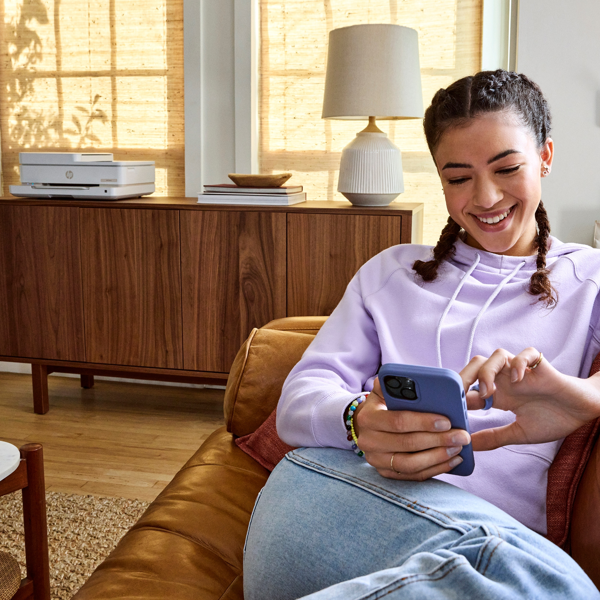 A woman with braids uses a phone on a brown leather couch. Printer and lamp in the background.