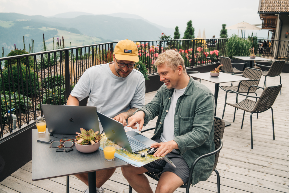 Dwóch mężczyzn z laptopami przy stole. Tło: góry, taras, widok.